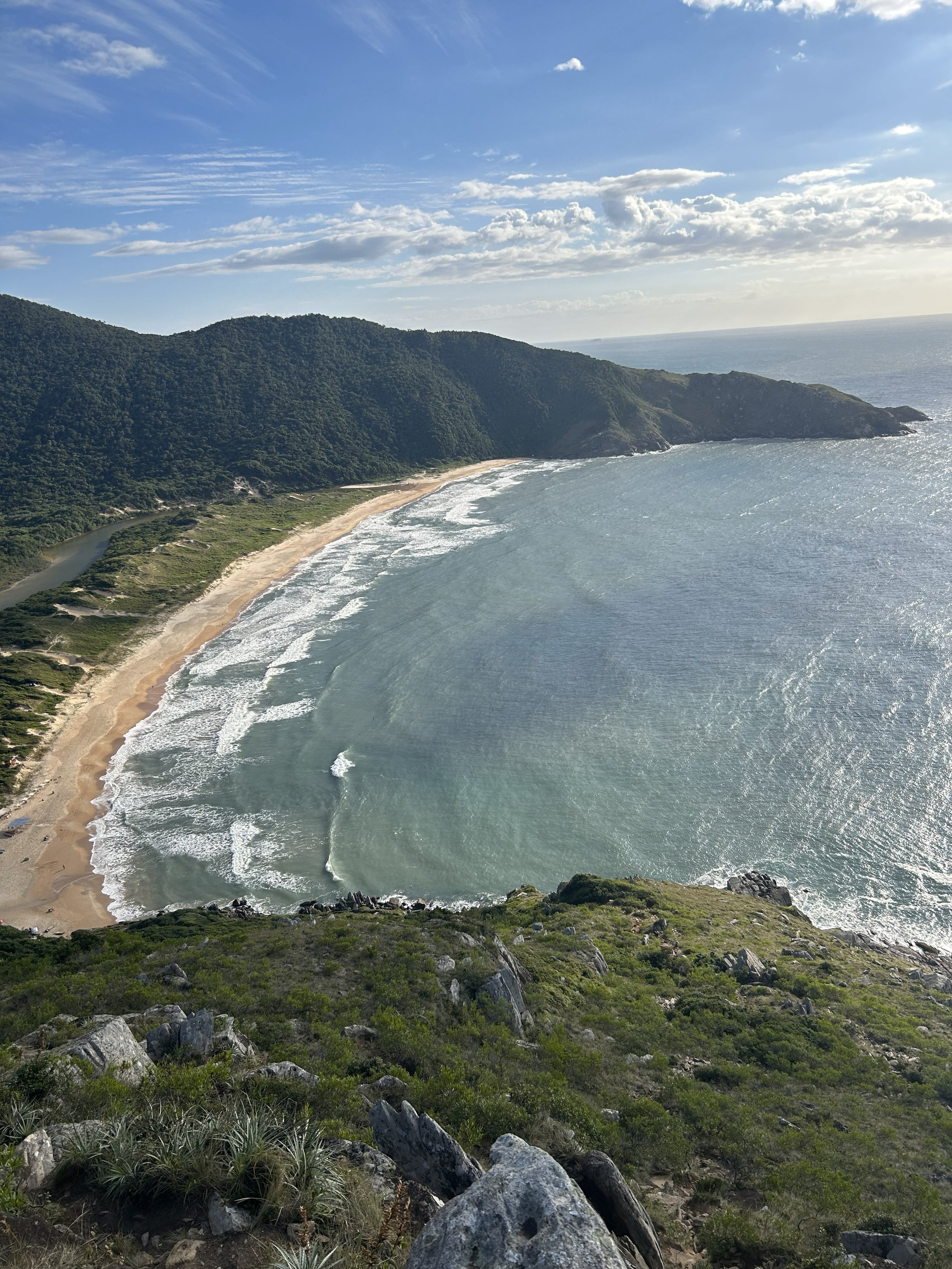 A scenic view from a hillside overlooking a crescent-shaped beach with rolling waves, green cliffs, and a partly cloudy sky. Lagoinha do Leste.