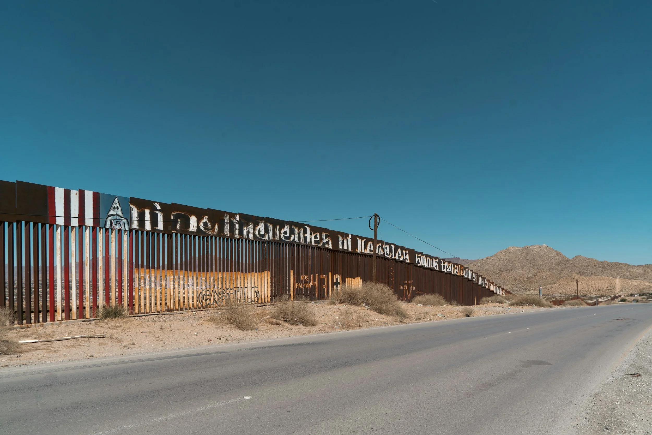 A long border fence with graffiti and painted signs, set in a desert landscape with mountains in the background and a clear blue sky.