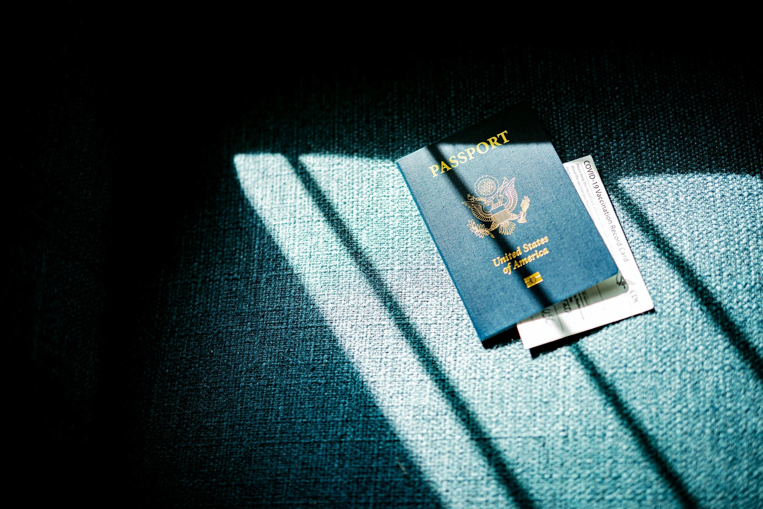 United States passport, vaccination record card, and boarding pass lying on a textured surface with sunlight casting shadows.