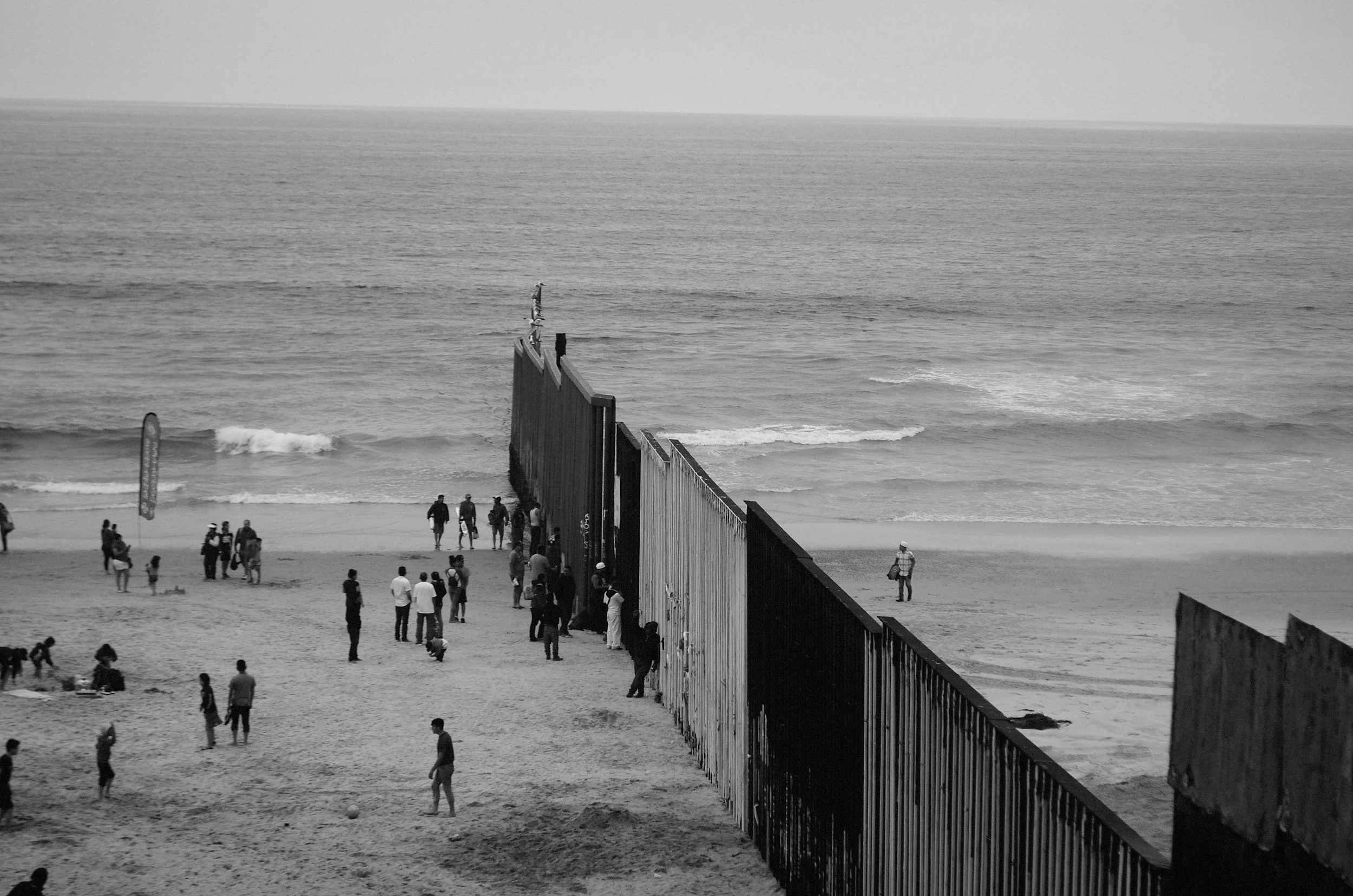 A black and white photo of the border wall extending into the ocean, with people on both sides and near the wall on the beach
