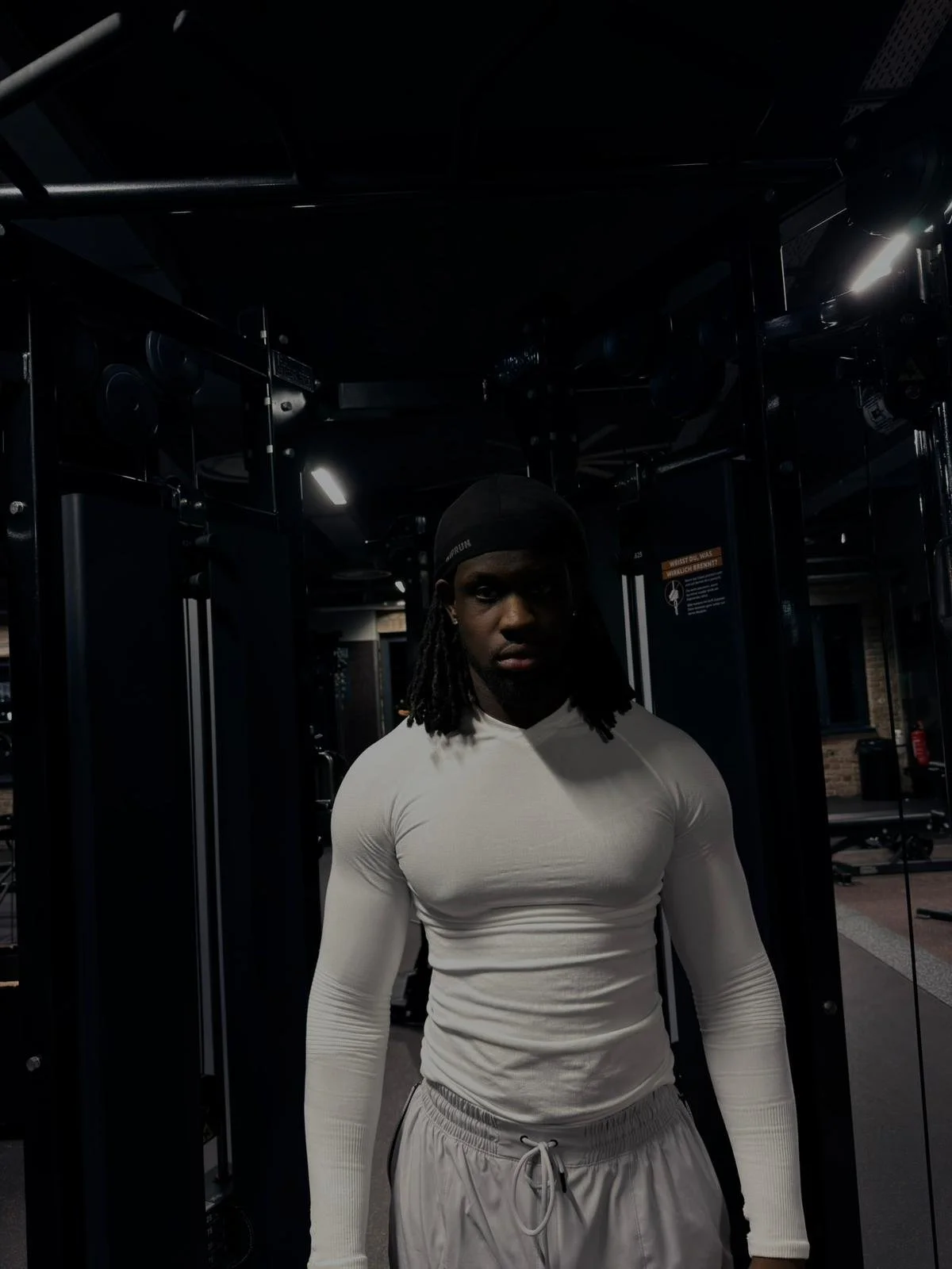Ein junger Mann in einem Fitnessstudio trägt ein langärmliges weißes Shirt, weiche Hosen und eine schwarze Mütze.