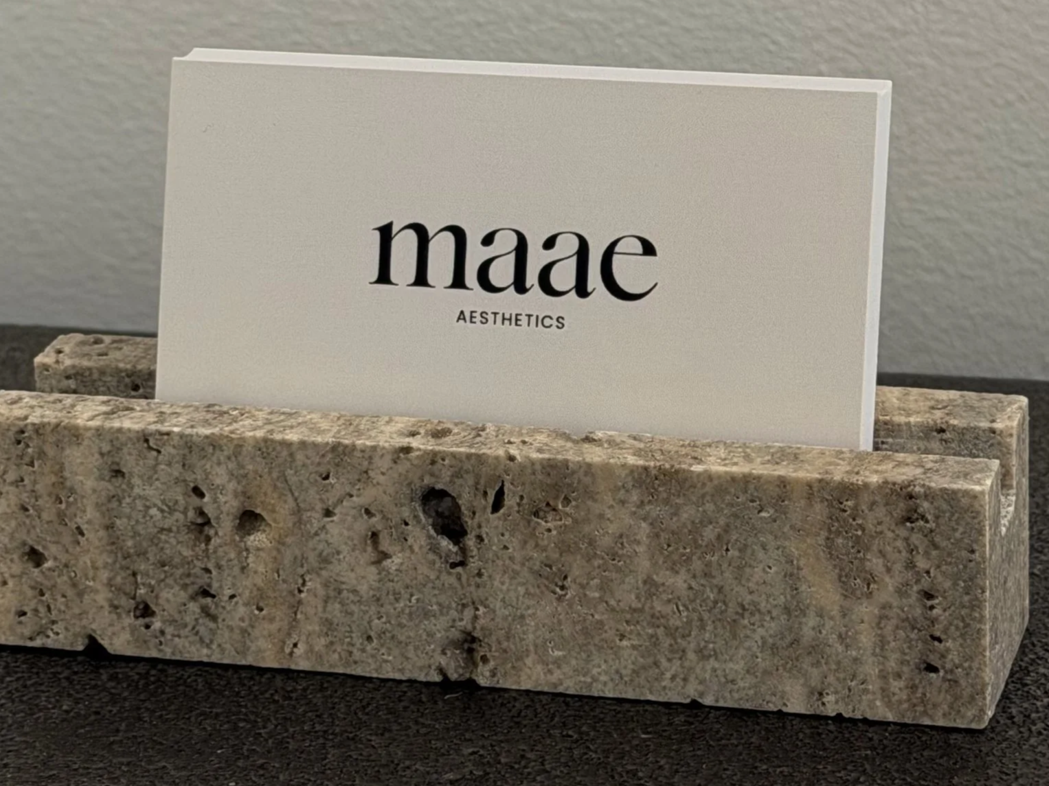 A business card for 'maae AESTHETICS' placed in a textured stone holder on a dark surface.