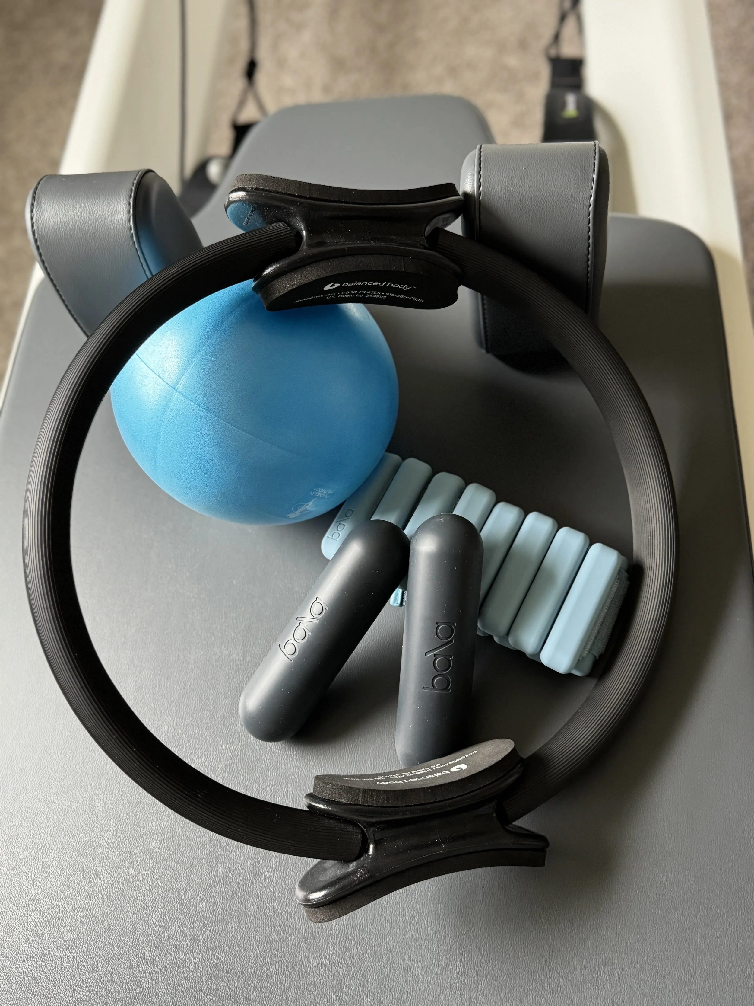 Fitness equipment including a blue exercise ball, a Pilates magic circle, two gray Bala Bars, a light blue Bala bangles weights, on a Balanced Body reformer .