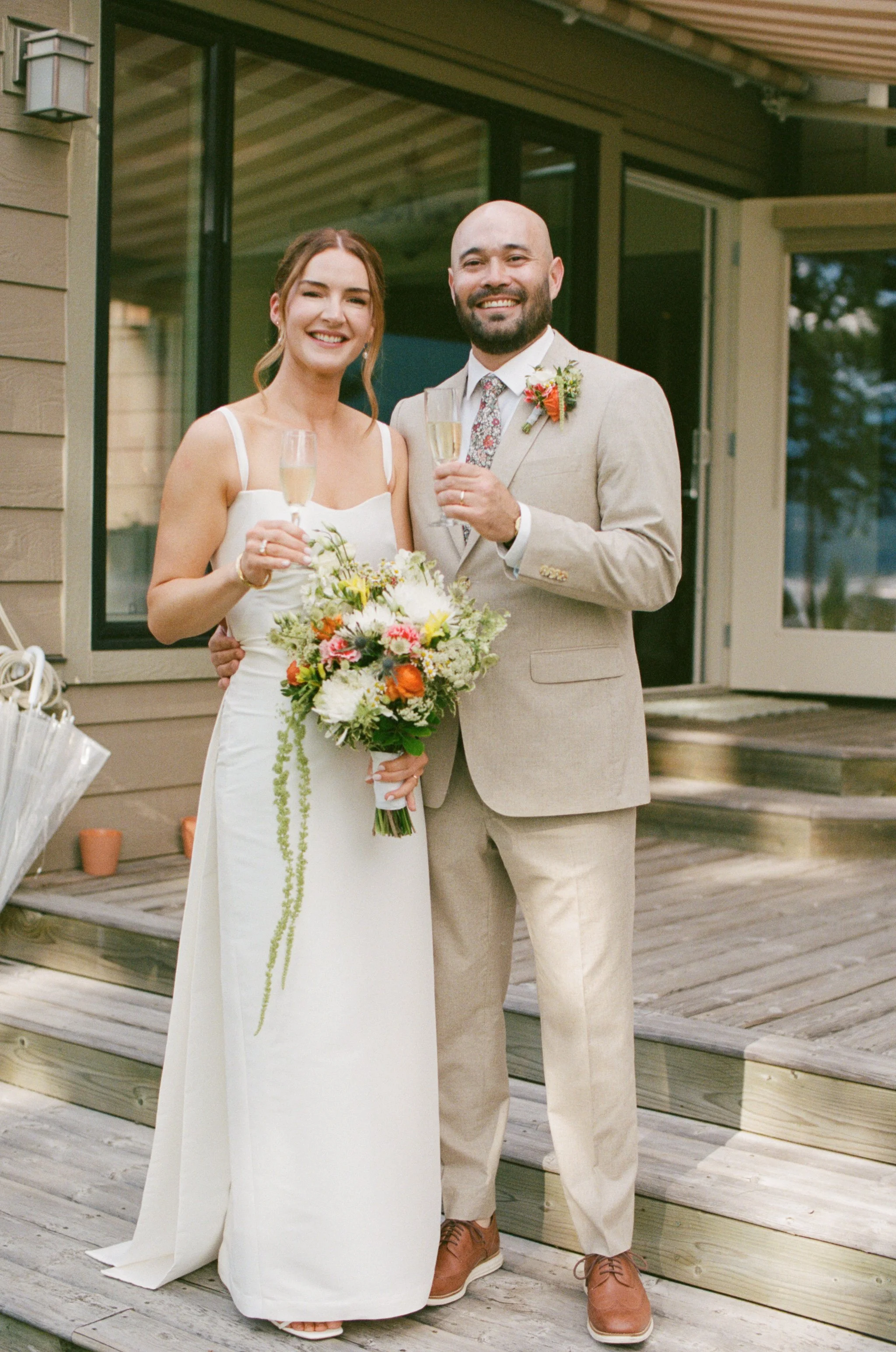 A bride and groom smiling and holding champagne glasses at their wedding, standing on a wooden deck outside a house, with the bride holding a bouquet of colorful flowers.
