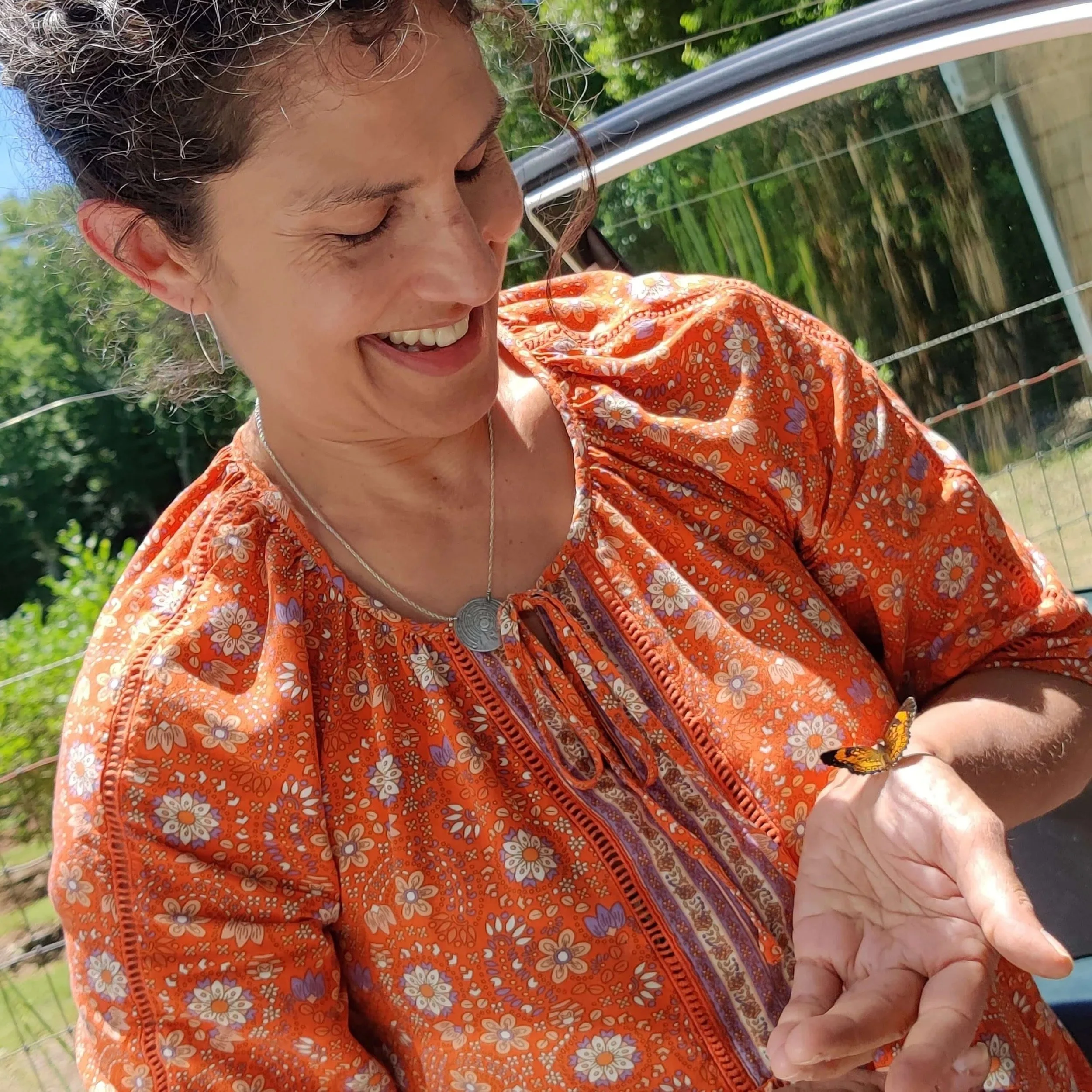 A woman with dark hair and earrings, wearing an orange floral blouse, is smiling as she holds a small butterfly on her hand outdoors on a sunny day.