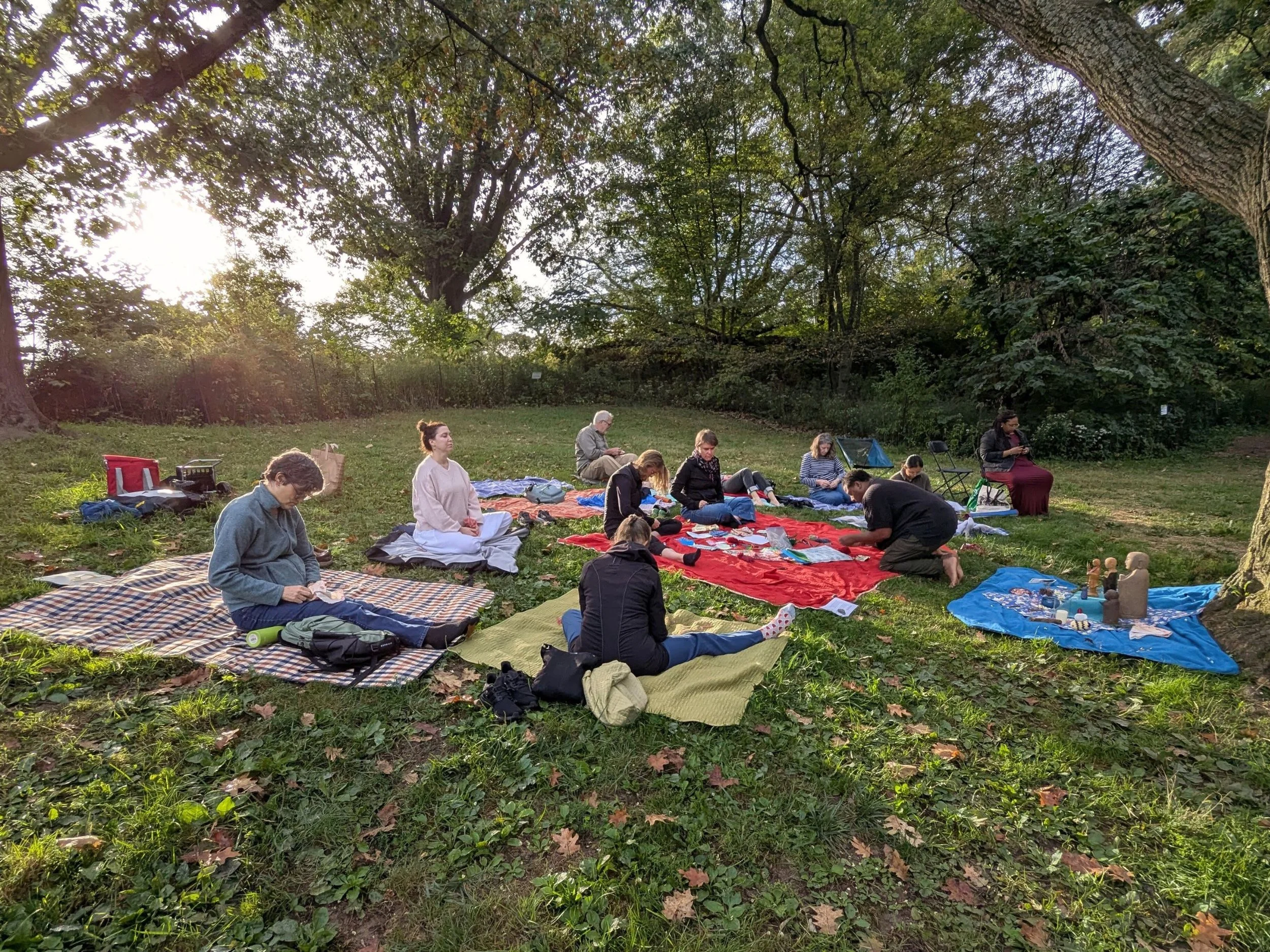 People sitting on blankets and mats outdoors under trees, participating in a spiritual or meditation gathering in a grassy park area.