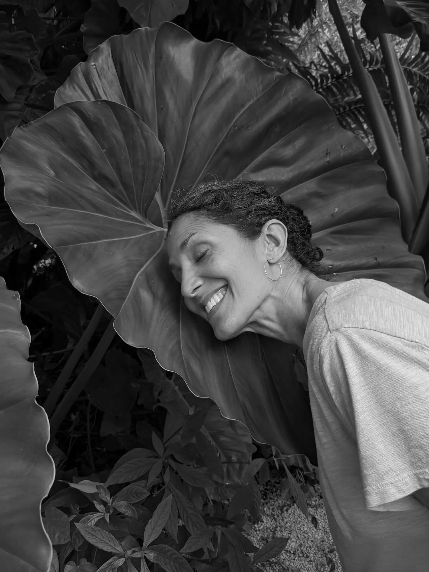 A woman with curly hair and hoop earrings smiling with her eyes closed, leaning her head against a large tropical leaf in a garden or jungle setting.