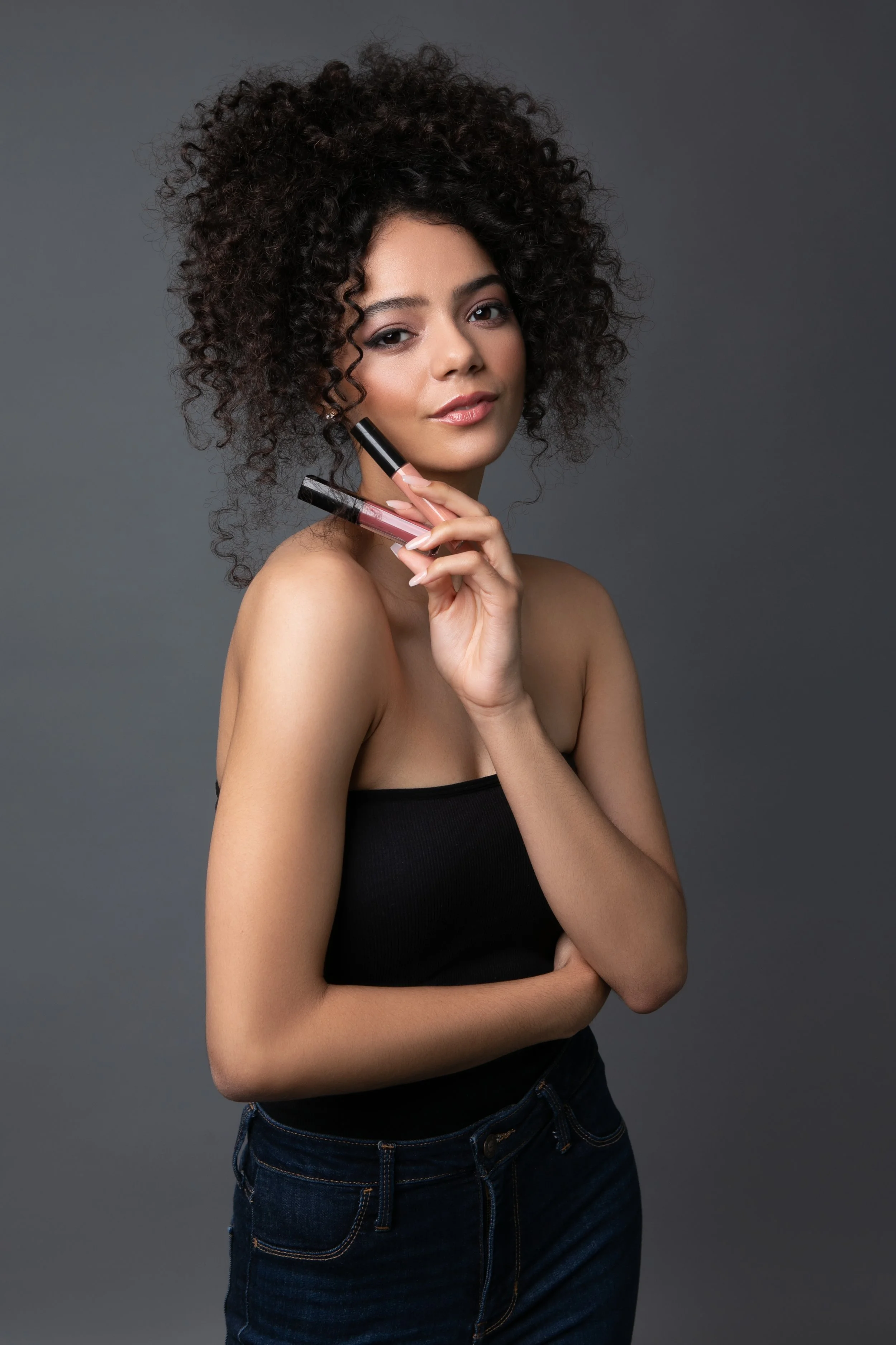 A woman with curly hair holding makeup lip glosses against a gray background.