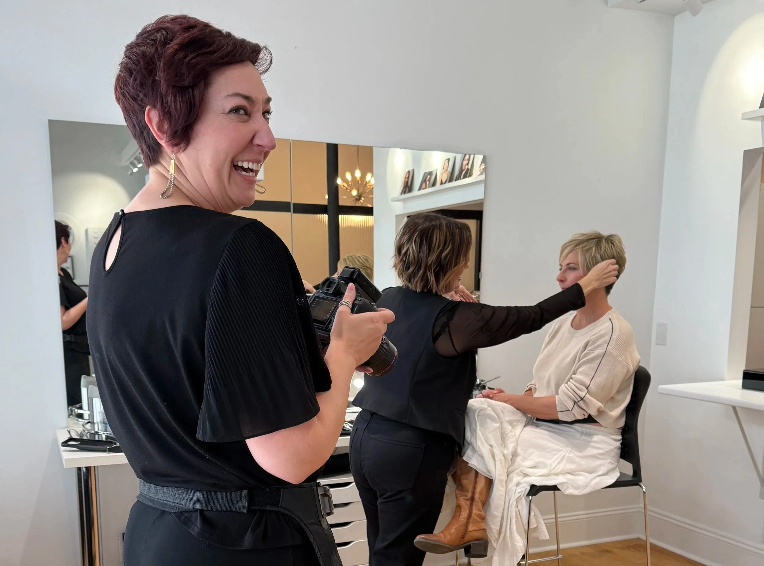 A hairstylist working on a woman sitting in a salon chair, while another woman holding a camera smiles in the foreground.