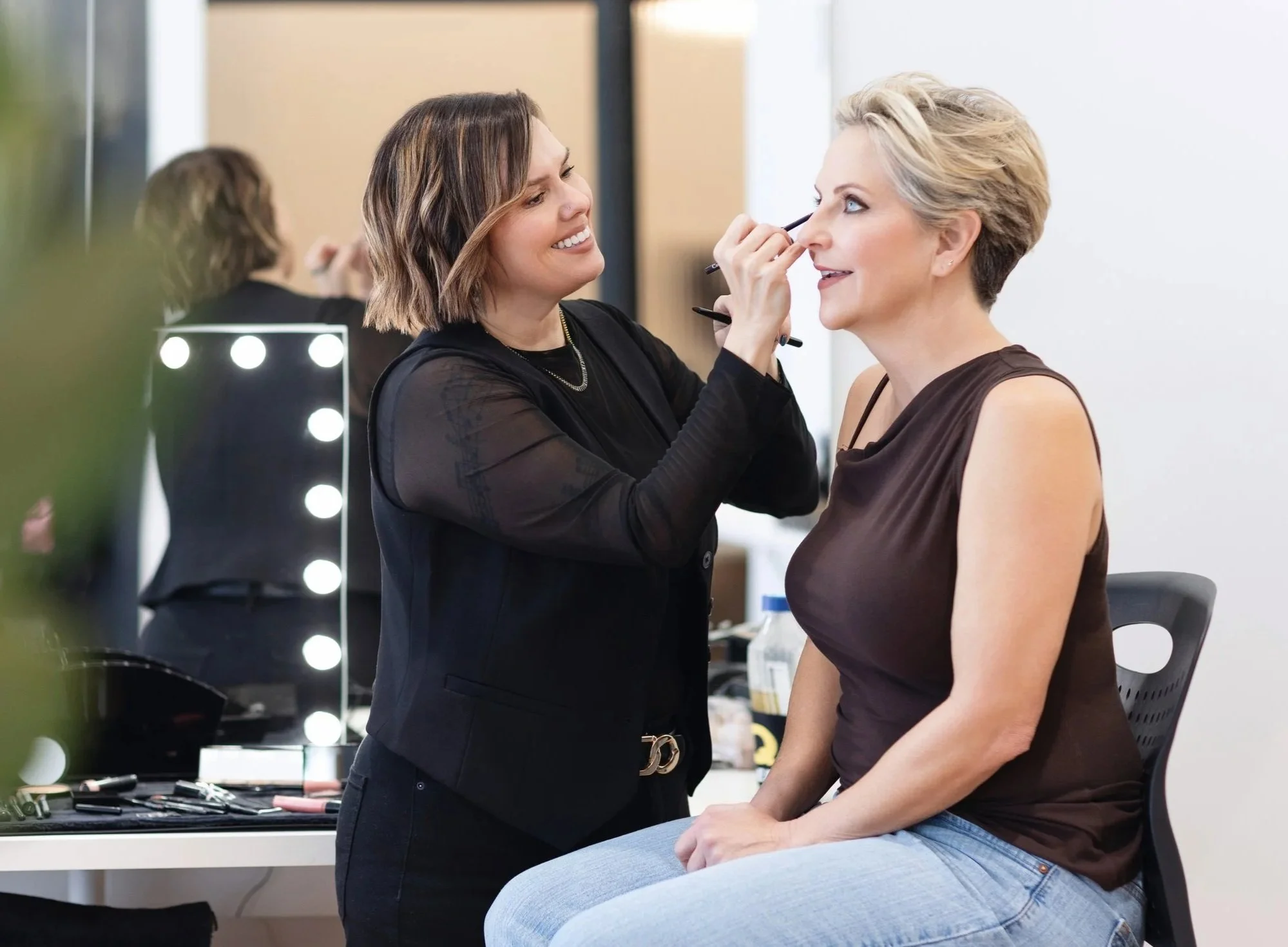 A professional makeup artist applies makeup to a woman sitting on a chair in a studio.