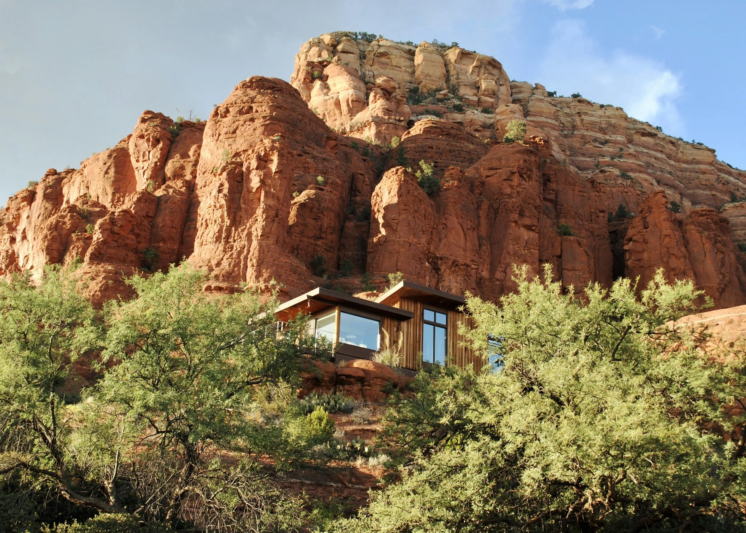 Modern house nestled among green trees at the base of tall red sandstone cliffs during daytime.