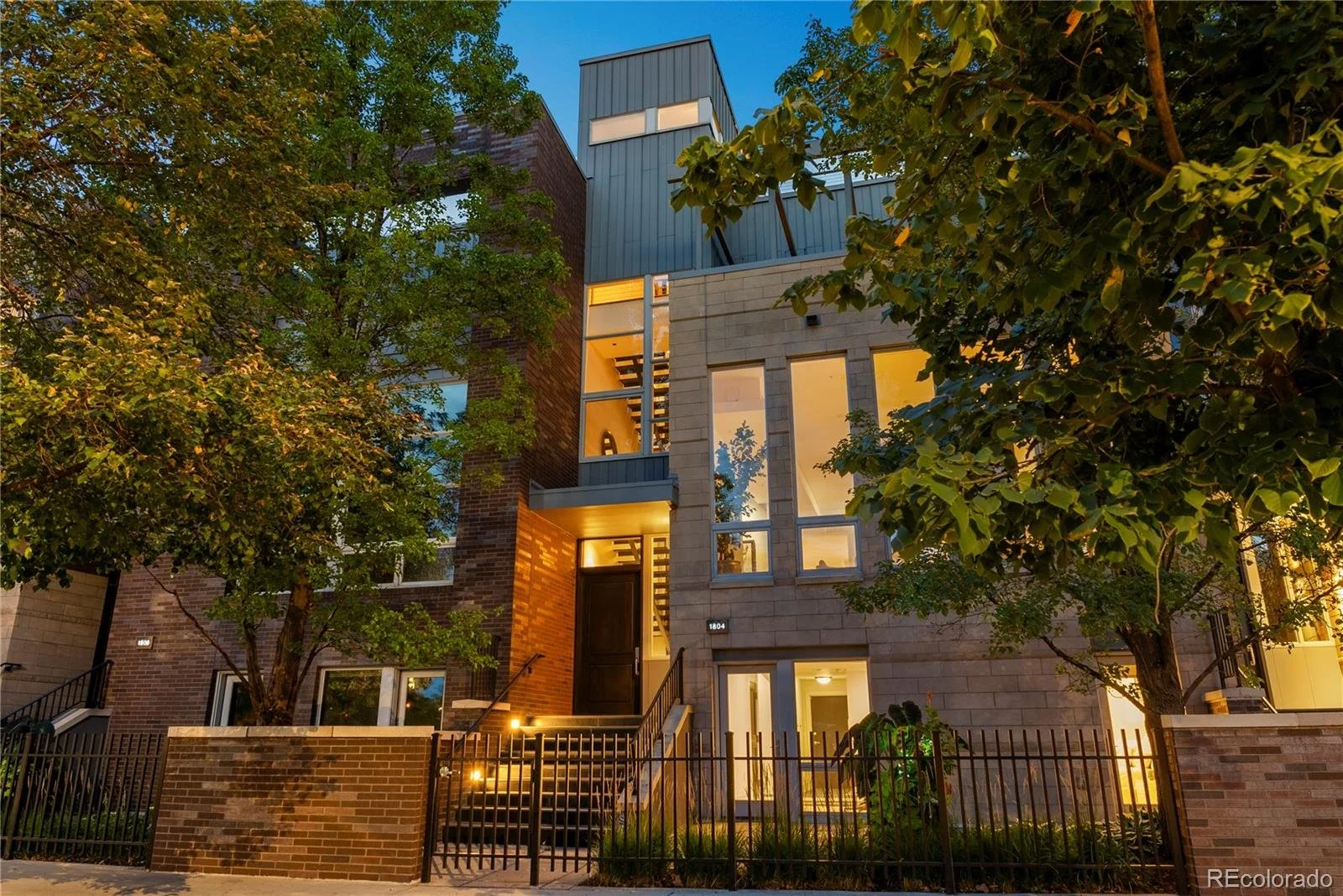 Modern multi-story house at dusk with large windows, trees, and a small front yard with a black fence.