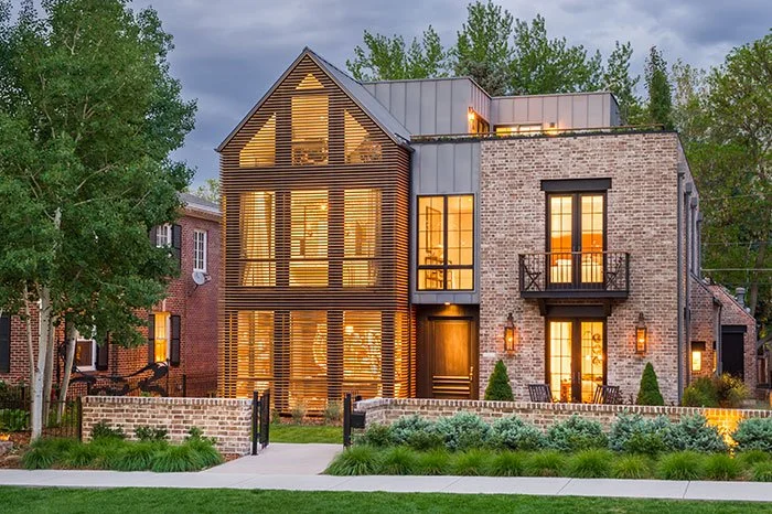 Modern multi-story house with large windows, brick and wood siding, and a small front yard with greenery.