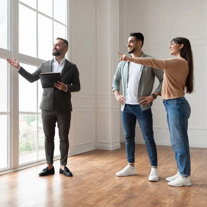 Three people standing in a bright room with large windows, engaging in a conversation. The person on the left, dressed in a suit, is holding a tablet and gesturing towards the window. The two others, a man and a woman, are smiling and pointing towards the window.