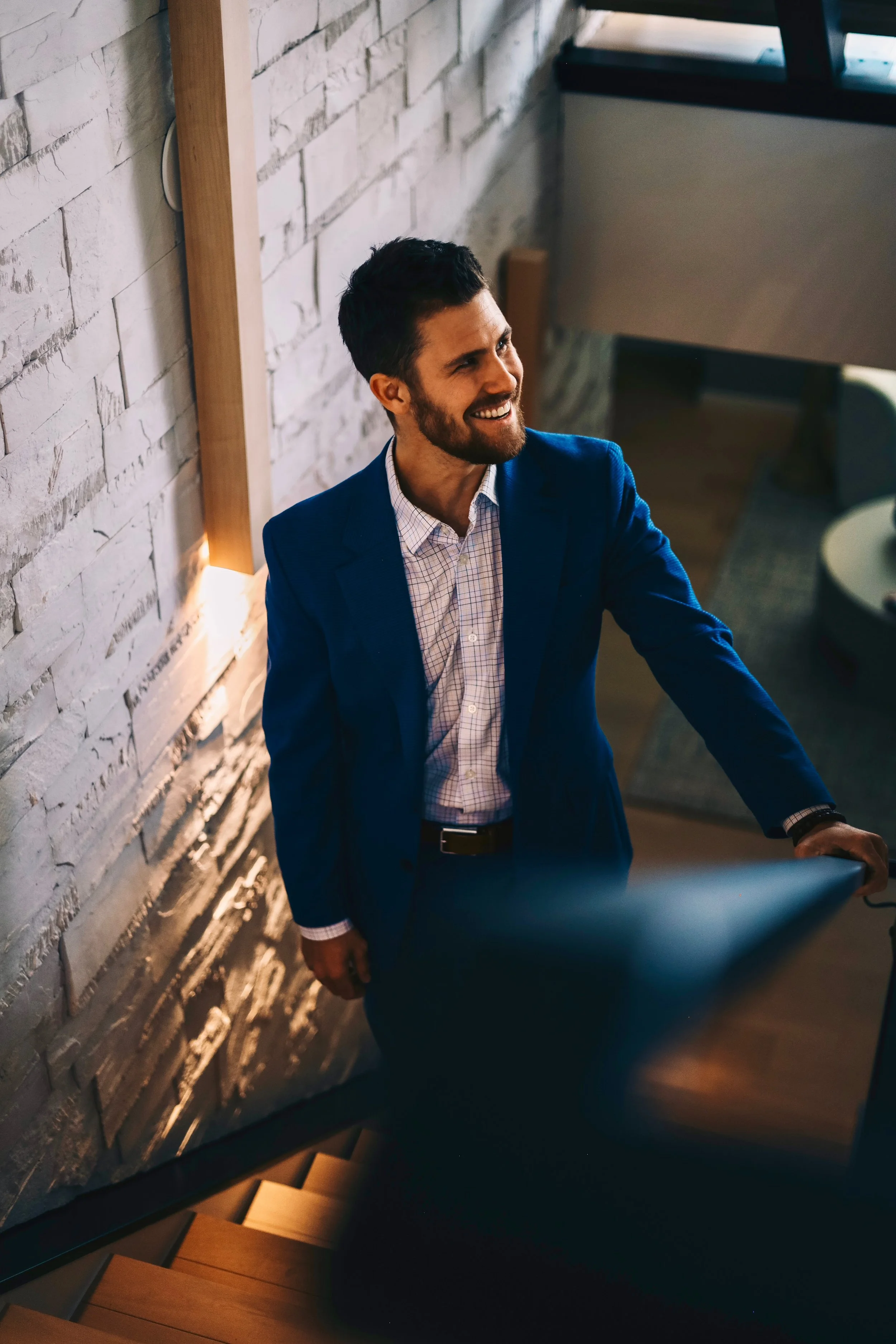 A man in a blue suit and white shirt standing on a staircase, smiling, next to a textured white brick wall indoors.