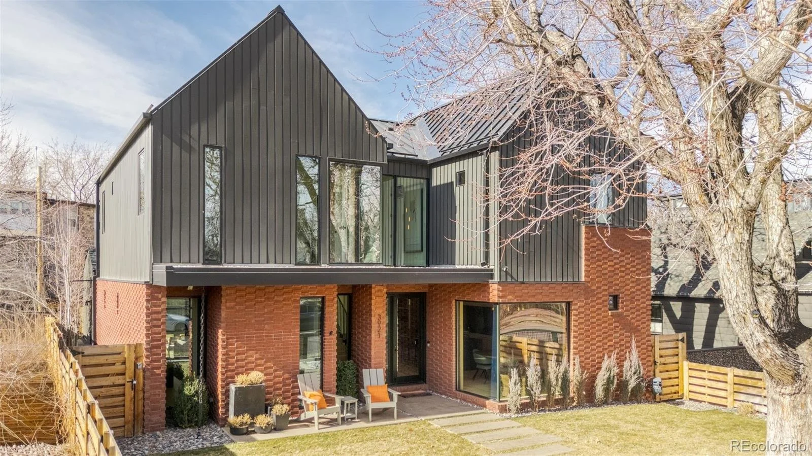 Modern two-story house with black vertical metal siding on the upper level and red brick on the lower level, large windows, outdoor patio with chairs and potted plants, and a large tree with bare branches in the yard.