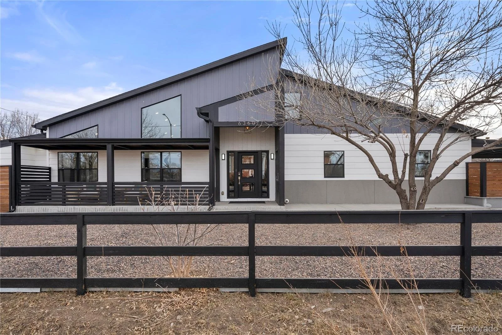 Modern house with dark gray and white exterior, large front door, and a leafless tree in front, enclosed by a black wooden fence, under a clear blue sky.