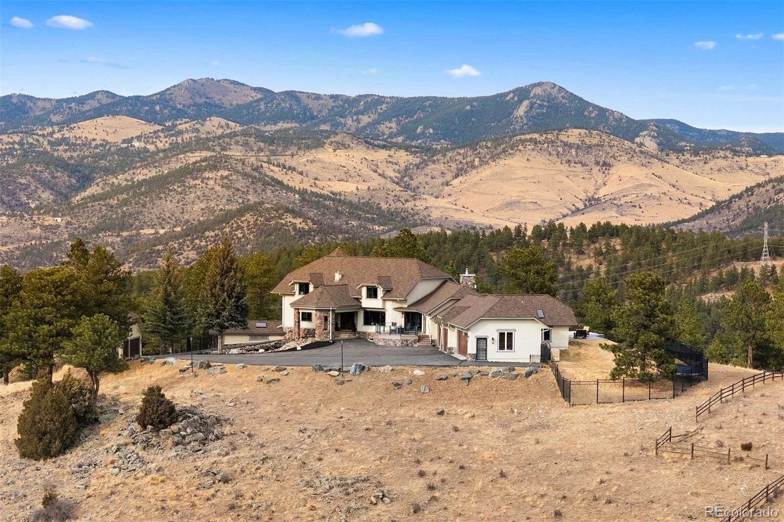 Large house with multiple sections and a brown roof, surrounded by trees and hills with mountains in the background under a blue sky.
