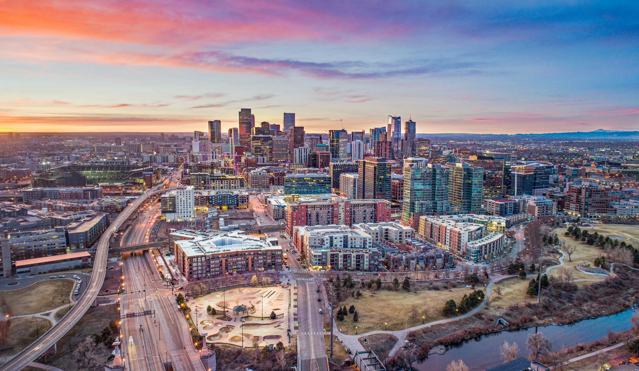 Aerial view of a city skyline during sunset with high-rise buildings, roads, parks, and a body of water.