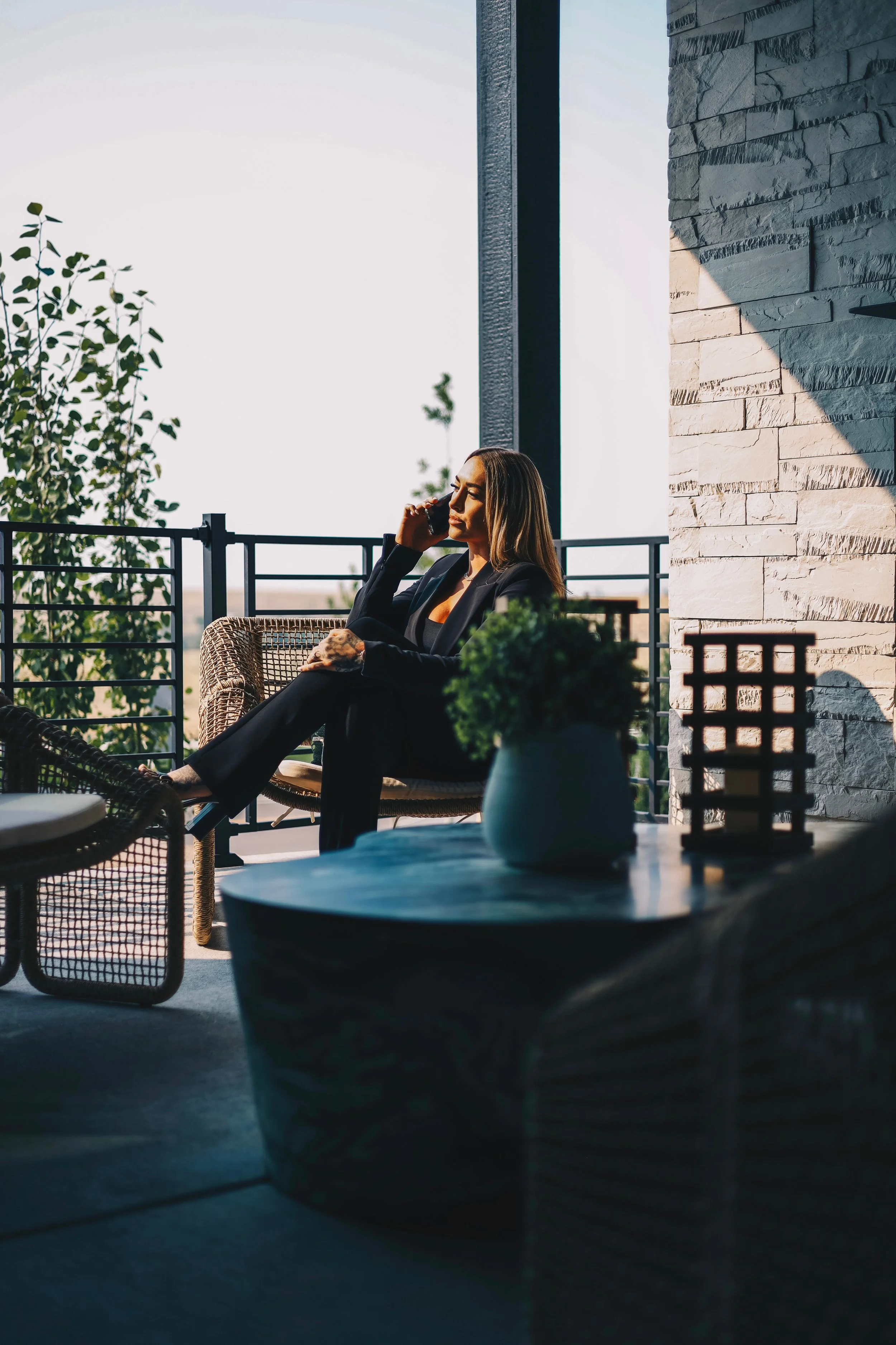 A woman in a black suit sitting on a balcony, talking on her cellphone, with a potted plant and decorative items on a table in the foreground.