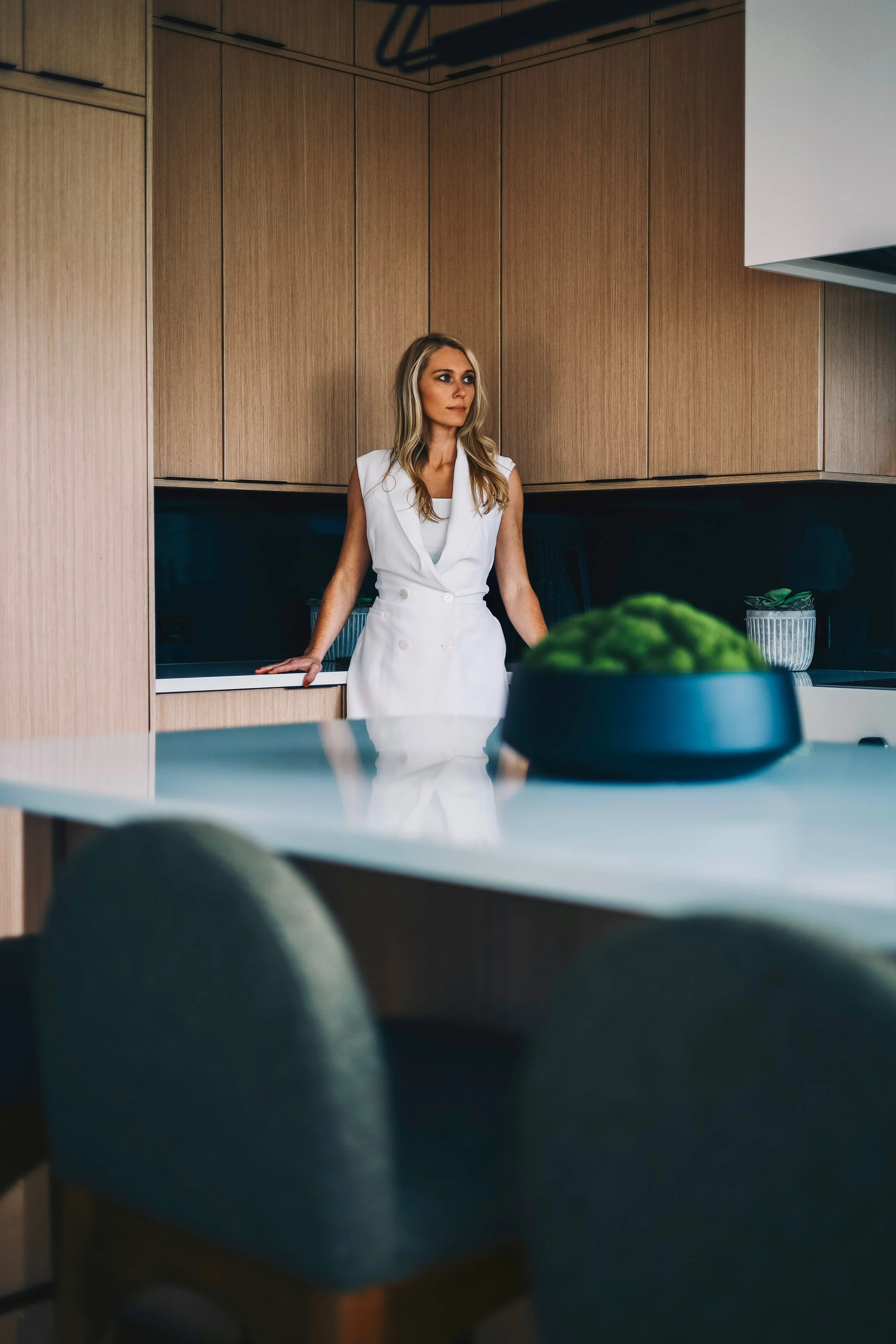 A woman with blonde hair wearing a sleeveless white dress standing in a modern kitchen with wooden cabinets, looking to her right while resting her hands on the counter.