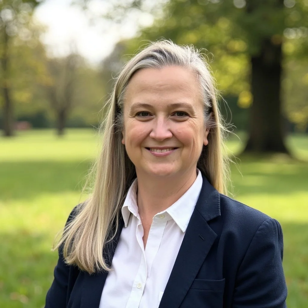 A woman with long blonde hair smiling outdoors in a park, wearing a white shirt and a dark blazer, with green trees and grass in the background.