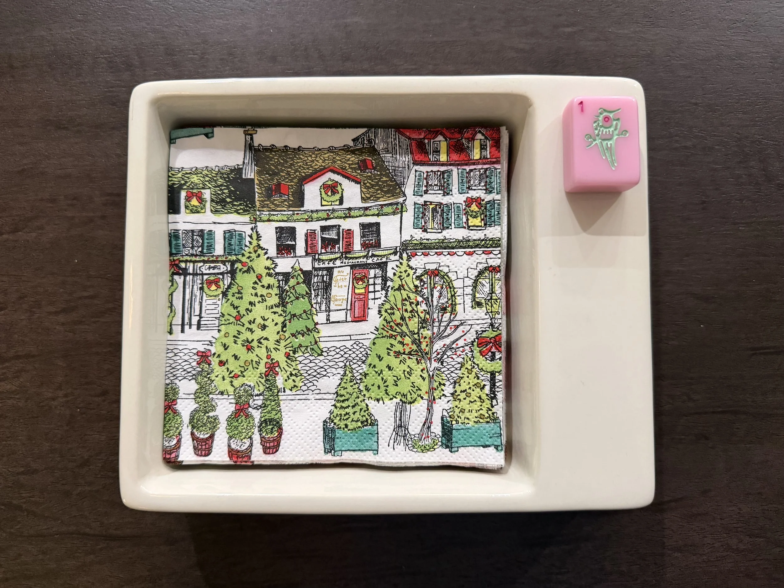 Decorative napkin with a Christmas-themed illustration of a festive street scene, including decorated trees, garlands, and buildings, inside a white game or display frame with a pink game piece featuring a spooky design in the top right corner.