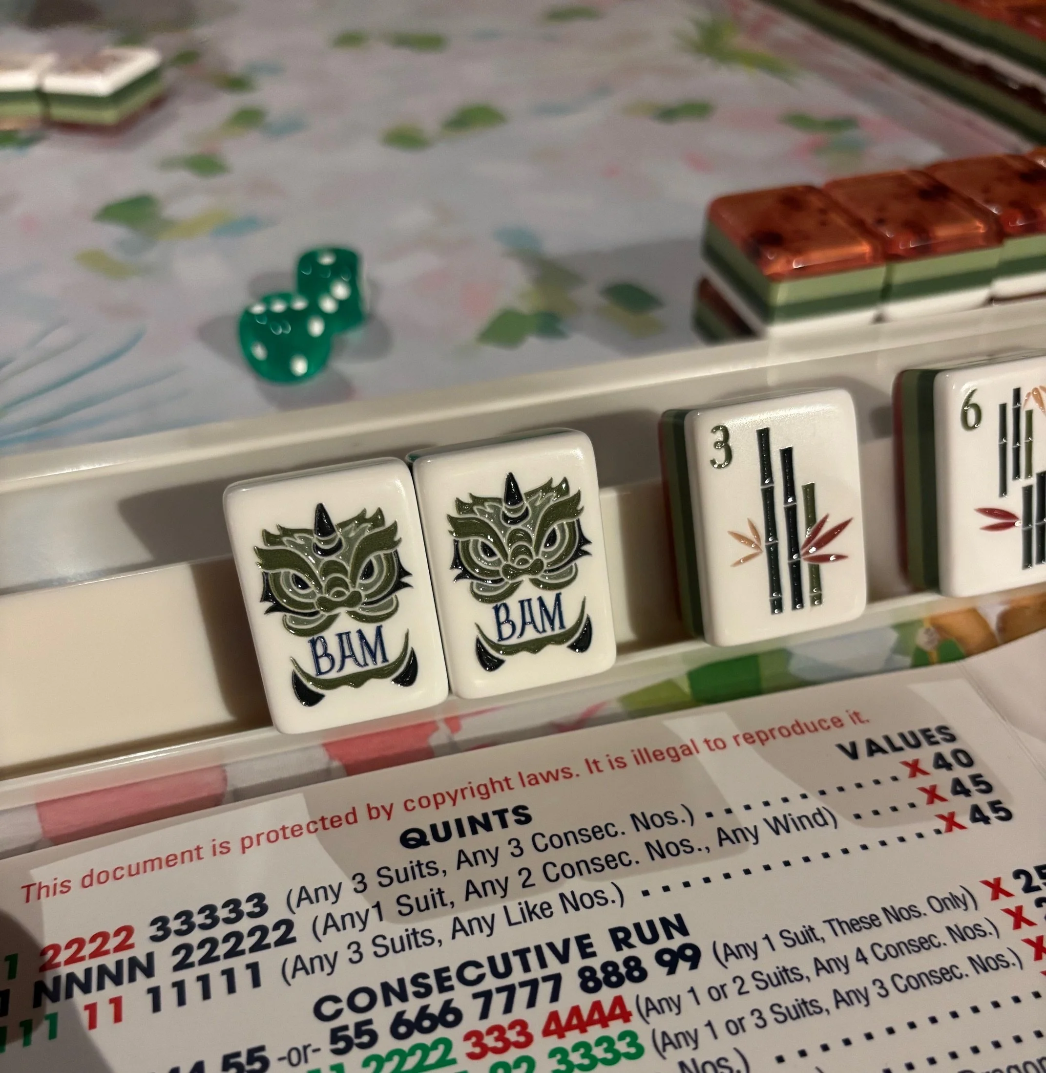 Close-up of Mah Jongg tiles showing two face-identifying tiles with a stylized face and the word 'BAM', and two column tiles with numbers 3 and 6. Part of the game setup with green dice and other tiles visible in the background.