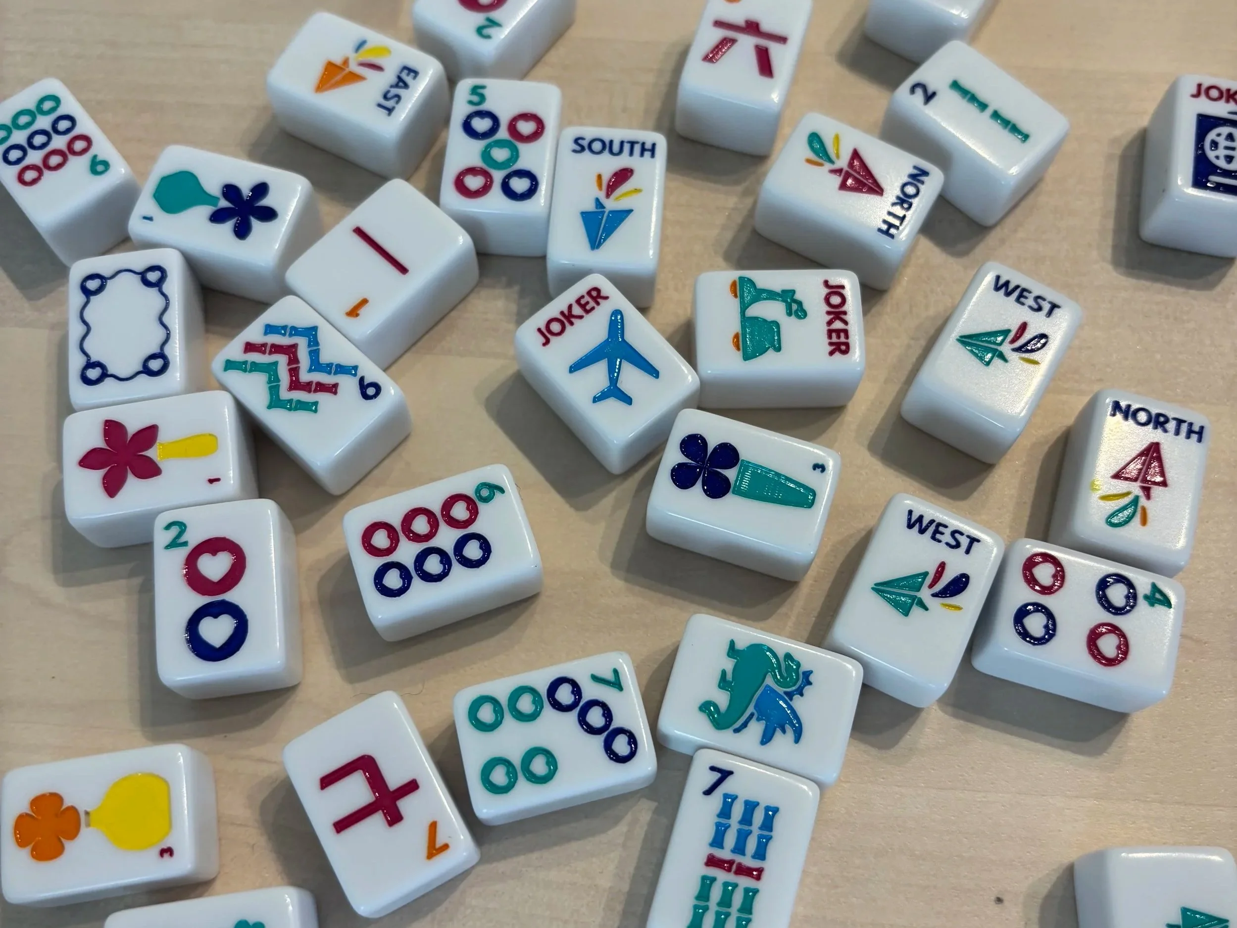 Close-up of scattered Mahjong tiles on a light wooden surface, displaying various suits and special tiles.