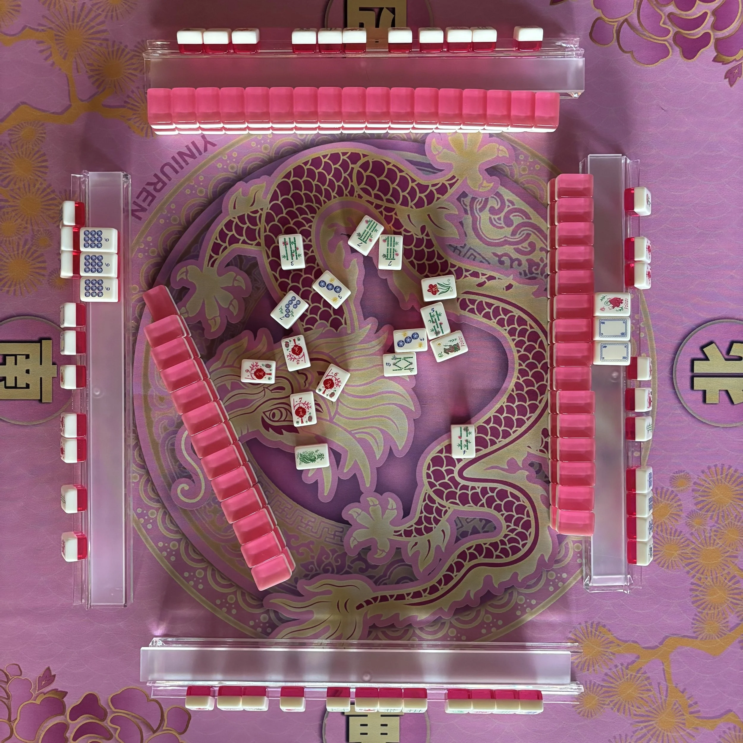 A Mahjong game in progress on a pink table with a dragon design. Various Mahjong tiles are arranged on the table, some face up and some face down. The tiles are decorated with Chinese characters, numbers, and symbols including flowers and dragons. There are four sets of tiles, each arranged in a row or group.