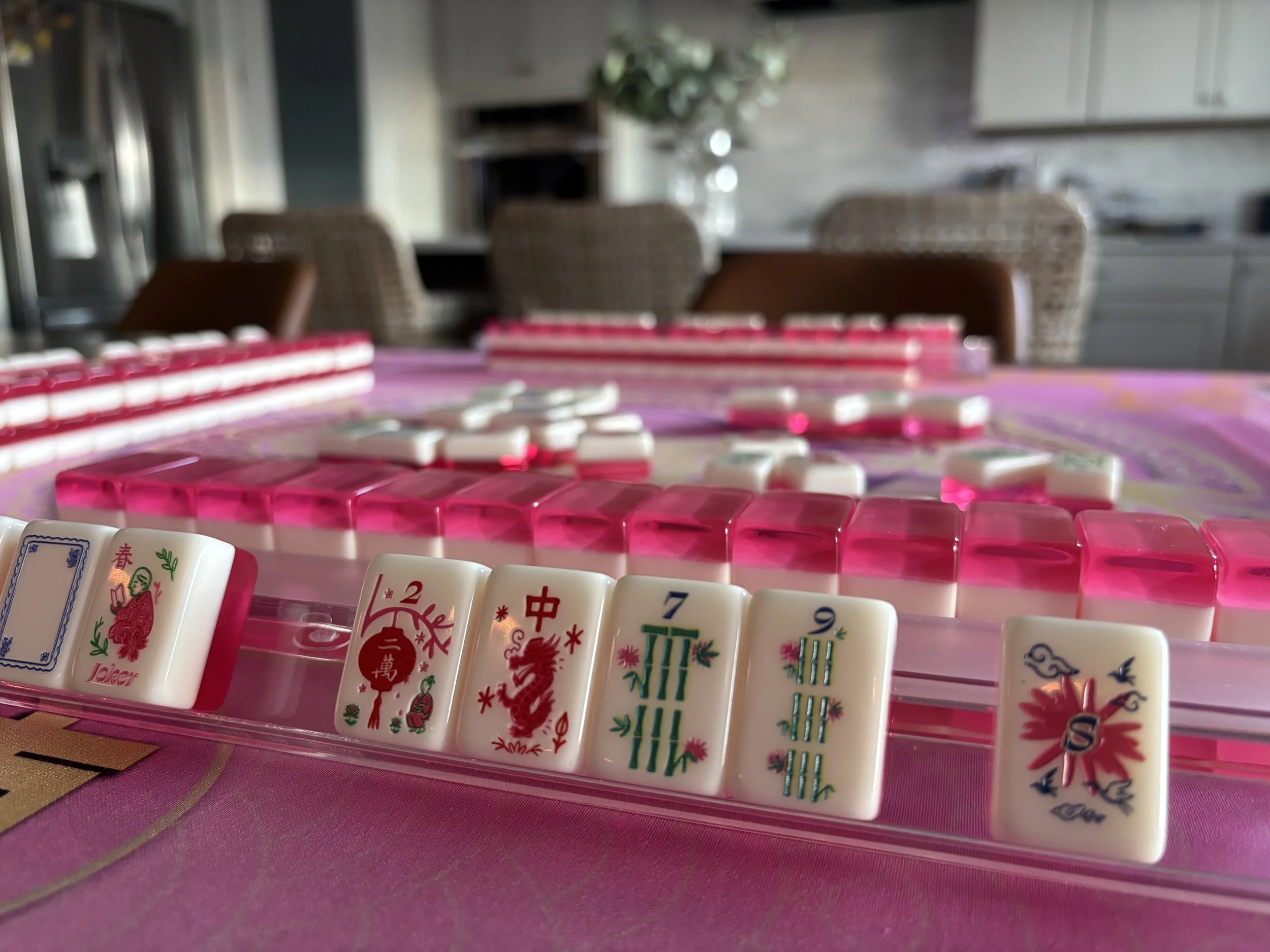 Close-up of mahjong tiles arranged on a pink tablecloth with a blurred background of a dining area.