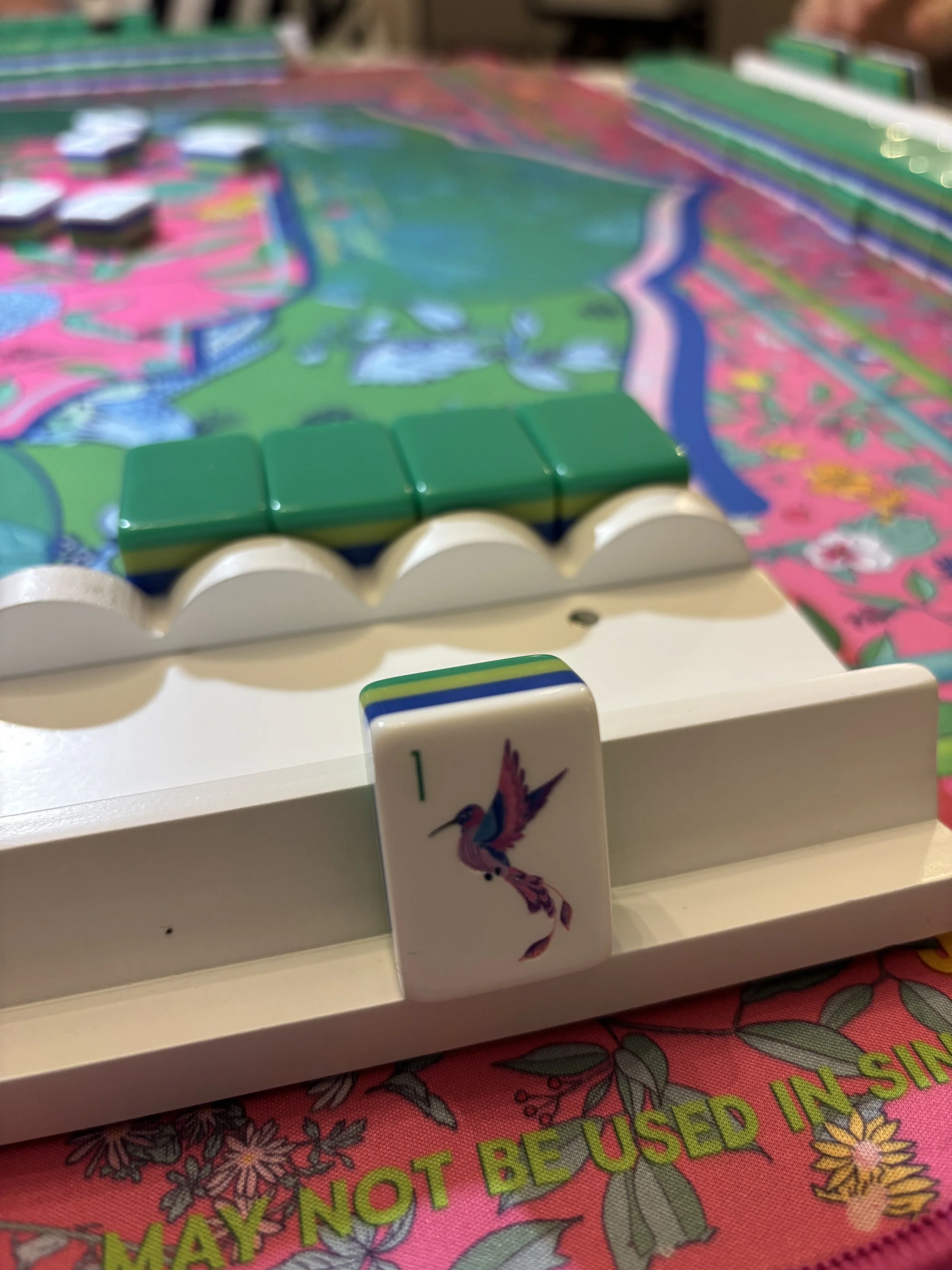 Close-up of a colorful board game with a white game piece featuring a hummingbird illustration and the number 1 on it. The game is set on a vibrant, floral-patterned surface with the words 'MAY NOT BE USED IN S' visible.