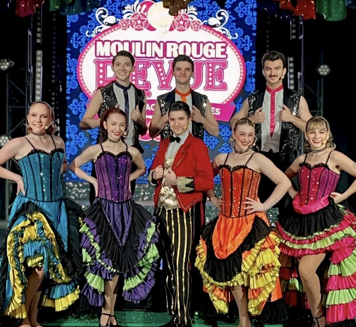 Group of performers in colorful costumes on stage with a Moulin Rouge revue sign in the background