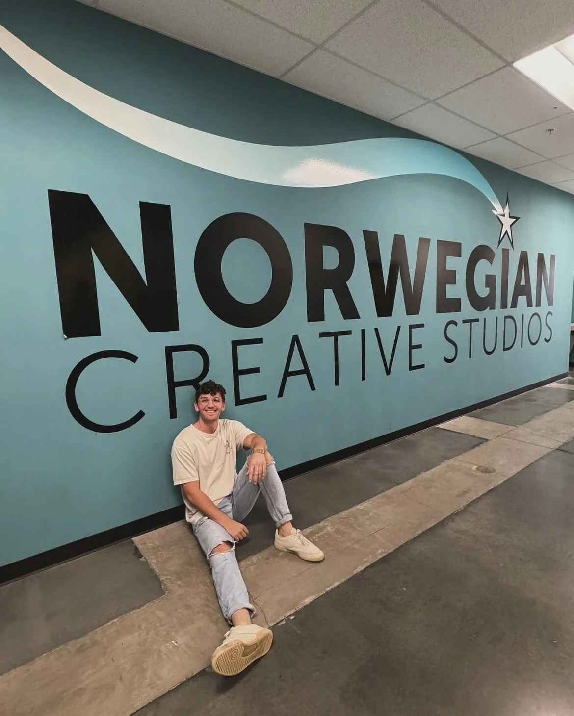 A young man sitting on the floor in front of a large wall mural that reads 'Norwegian Creative Studios' with a shooting star graphic.