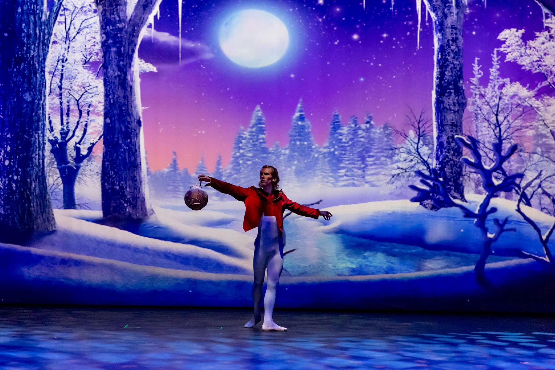 Ballet dancer in a red jacket, white tights, and ballet shoes performing on stage with a digital winter landscape background showing snow-covered trees, a full moon, and a starry sky.