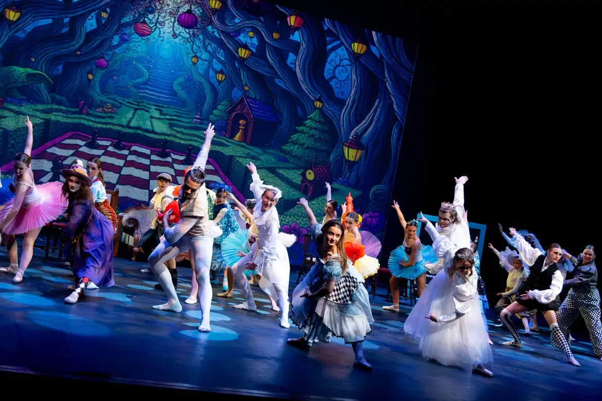 Children performing a ballet on stage with a colorful, fairy-tale forest backdrop, dressed in costumes like tutus, dresses, and whimsical outfits.