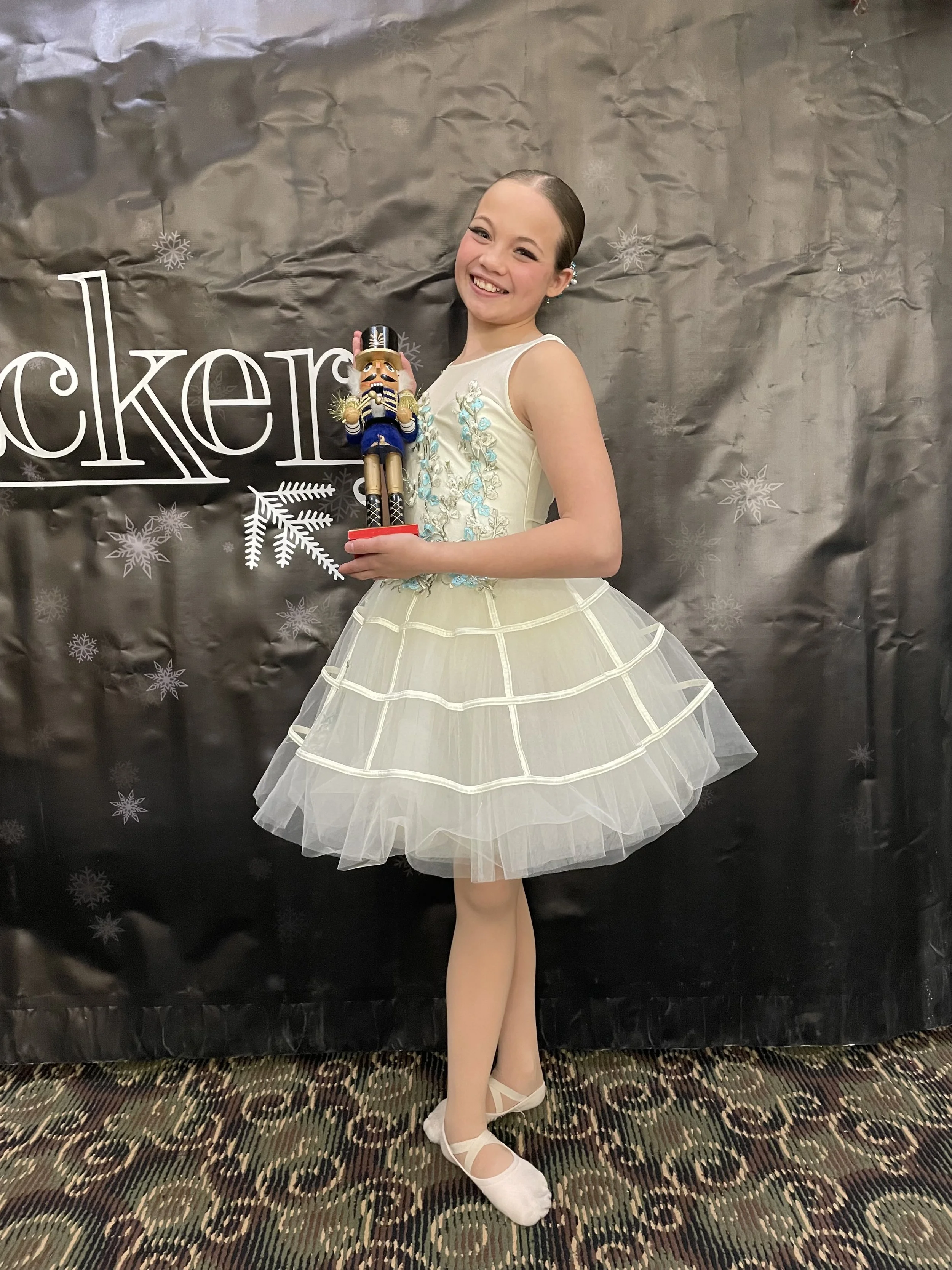 A young girl in a cream-colored ballet dress with blue embroidery, holding a nutcracker figurine, standing on a decorated stage with a black backdrop with snowflakes and the word 'rockets'.