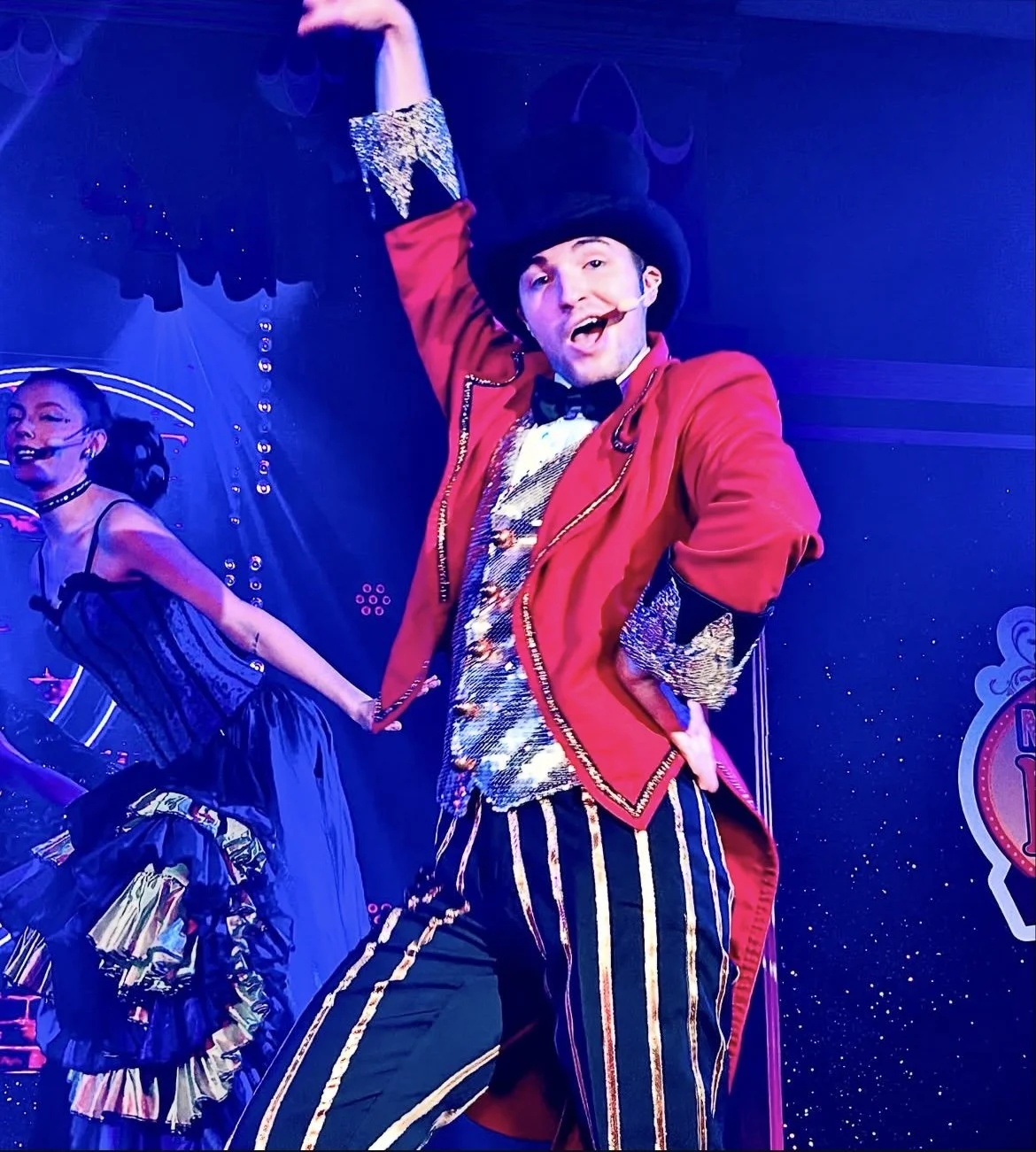 Performer wearing a red tailcoat, striped pants, and a black hat, singing with a microphone headset on, on a stage with vibrant lighting.