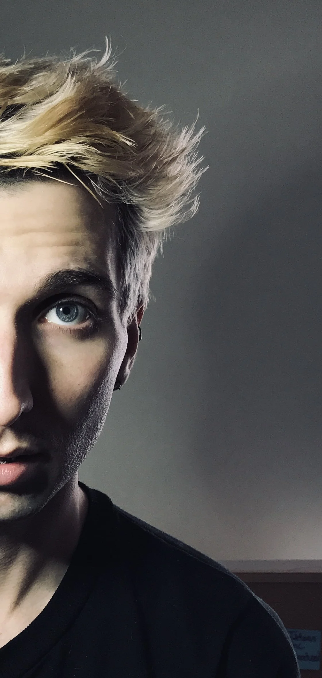 Half of a person's face with blonde hair, blue eye, and ear visible, wearing a black shirt, against a dark background.