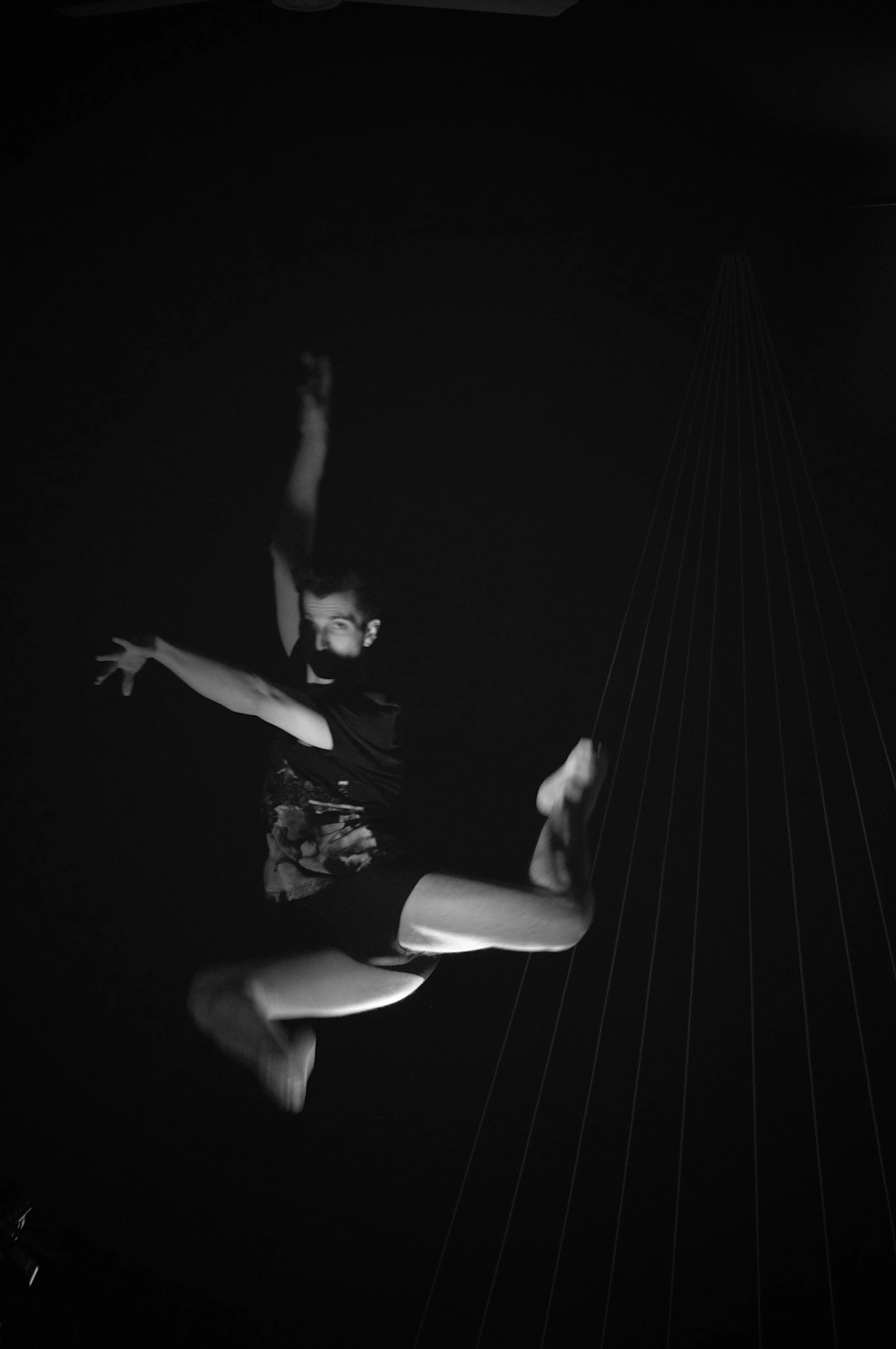 A person performing an aerial dance move, mid-air, in a dark setting, illuminated by a light source. The person is wearing shorts and a T-shirt, with their arms and legs extended dynamically.