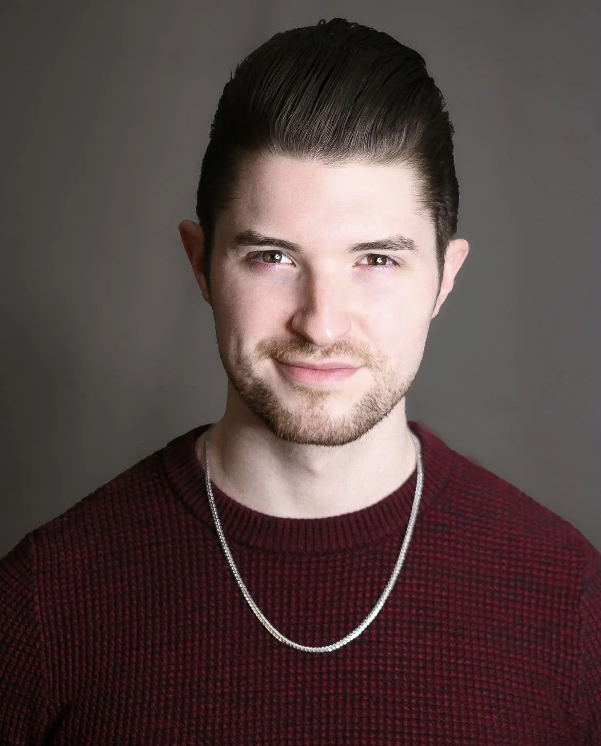 Young man with dark hair and light skin, wearing a red sweater and a silver chain necklace, smiling against a neutral gray background.