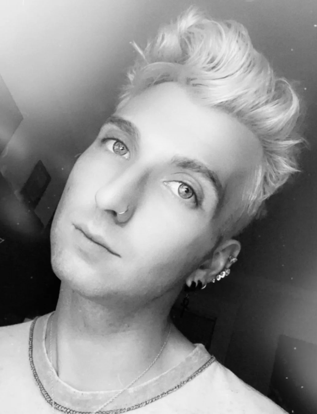 A black and white close-up portrait of a young person with short, textured hair, piercing eyes, multiple earrings, and a small nose piercing.