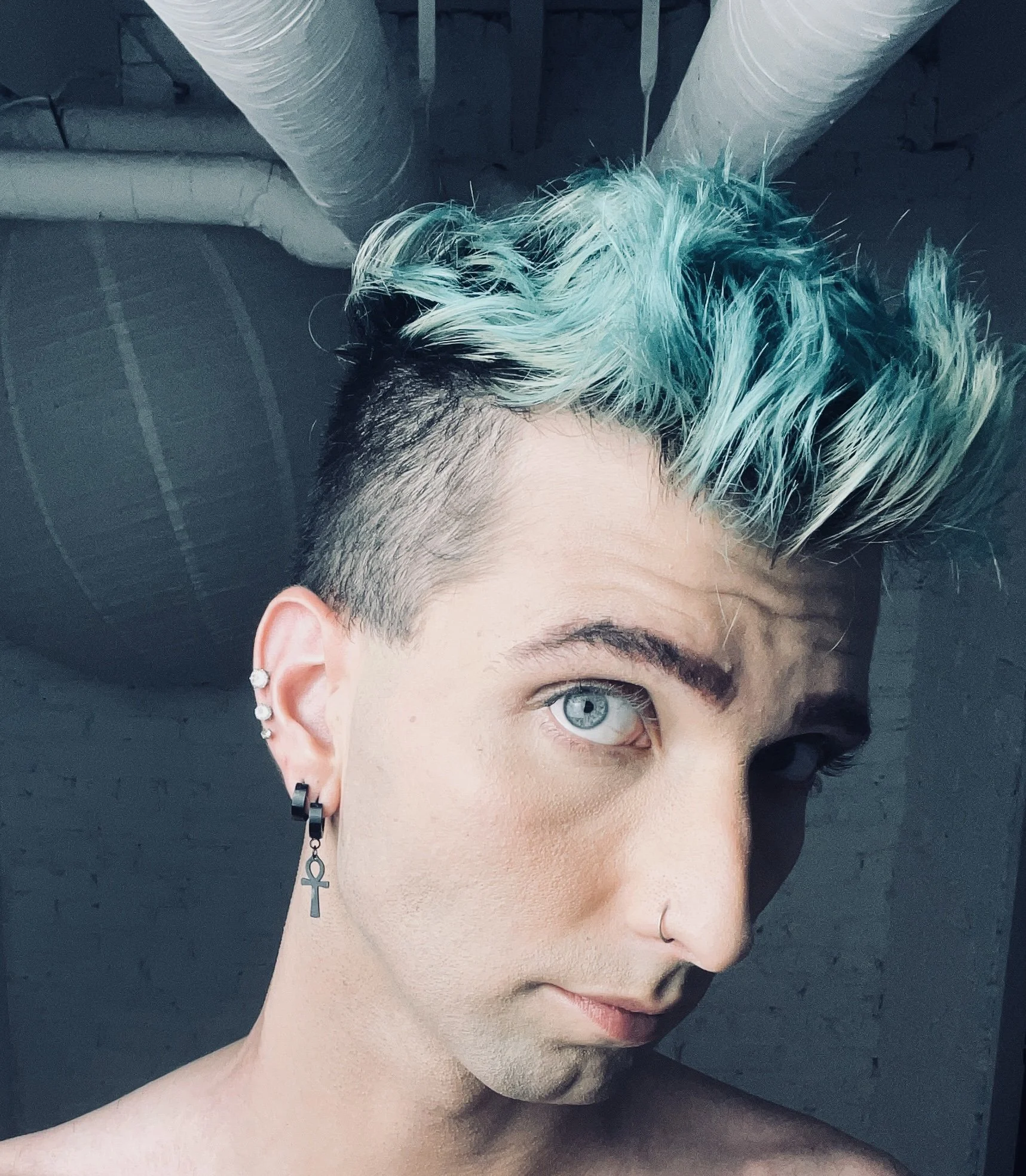A person with short, black hair and dyed voluminous, teal-colored hair on top, with a side shave, wearing multiple earrings including an ankh pendant, and a nose ring, taking a selfie in a room with exposed ductwork and brick wall.