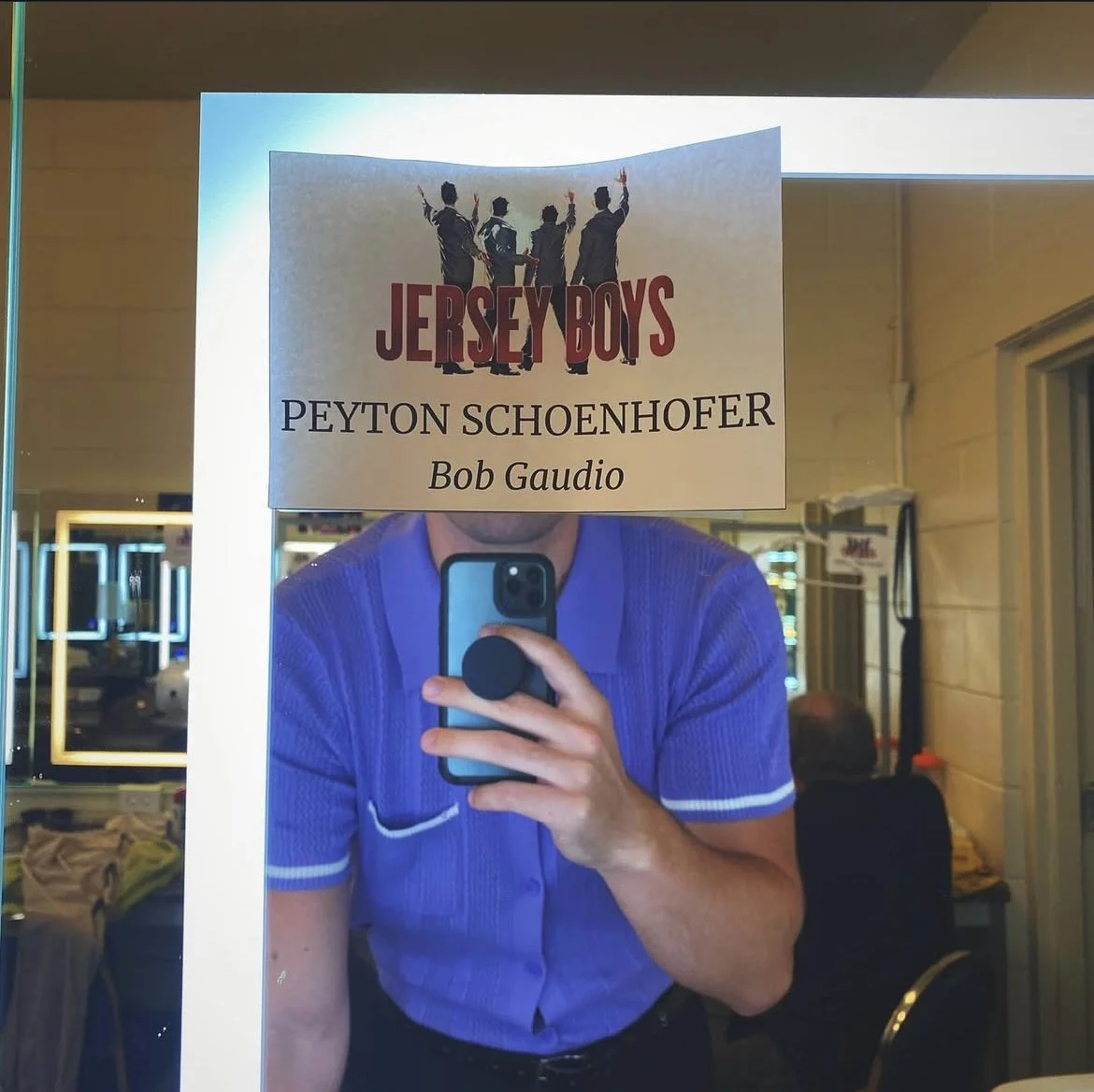 Mirror reflection of a person taking a photo with a phone, holding a sign for the Jersey Boys musical featuring Peyton Schoenhofer and Bob Gaudio, with the person wearing a blue polo shirt.