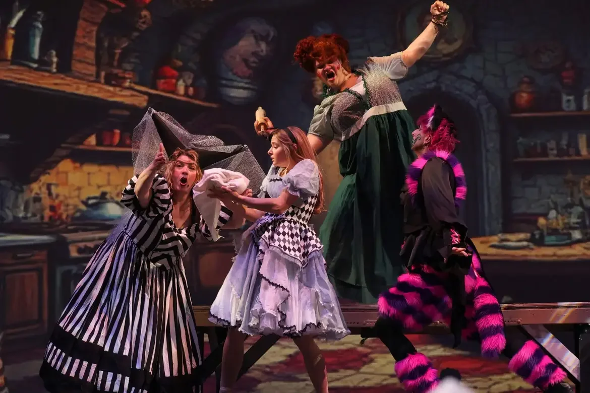 A theatrical scene with four actors in colorful costumes, appearing dramatic and expressive, set against a painted rustic kitchen background.