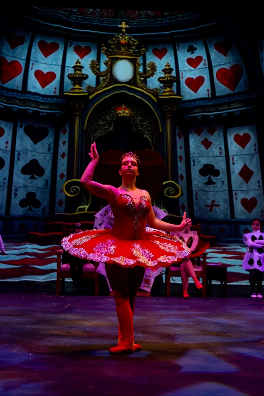 A ballet dancer in a red tutu with a tiara performs on stage in front of a fantasy castle backdrop decorated with playing card suits. Other dancers are seated in the background.