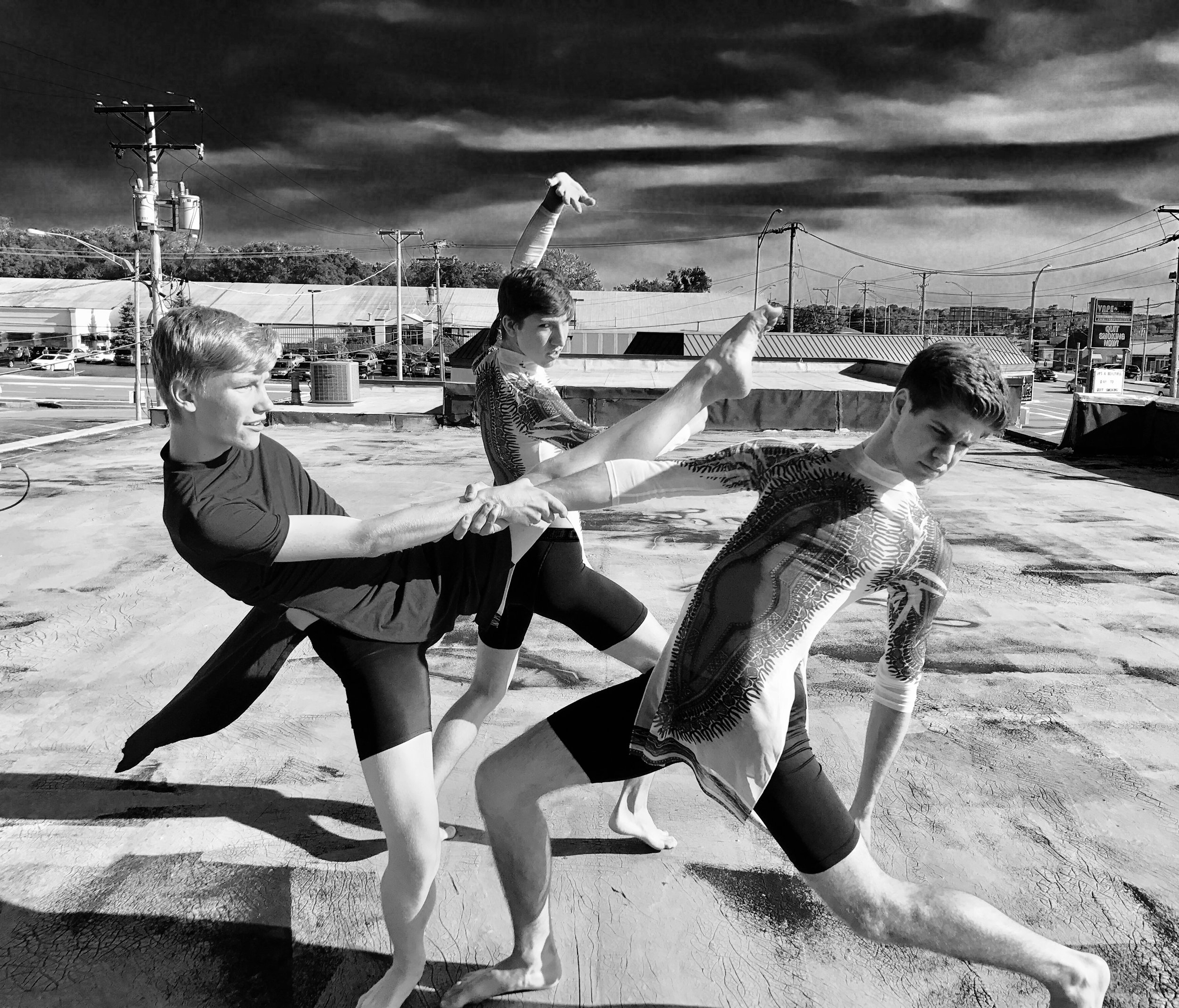 Three young individuals practicing martial arts or dance on a rooftop, with a cloudy sky and urban landscape in the background.
