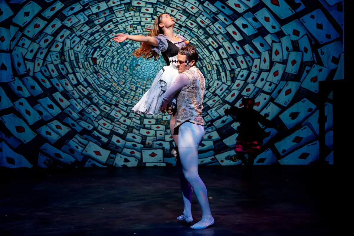Two ballet dancers perform on stage in front of a background with a swirling pattern of playing cards, with one dancer holding the other in a lifted pose.
