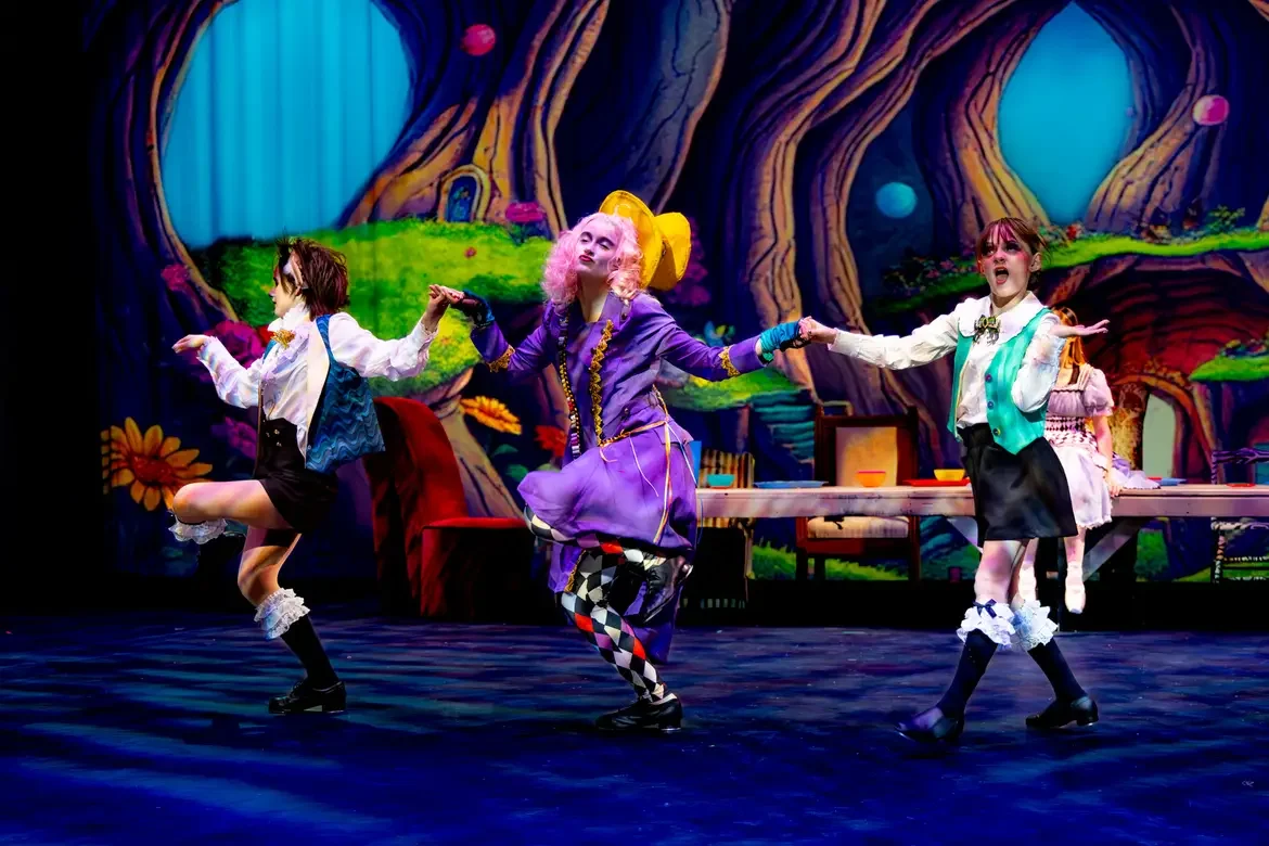 Performers in a colorful theatrical production, with a woman in a purple costume and three girls in vintage-style outfits, on a stage with a vibrant fantasy forest backdrop.