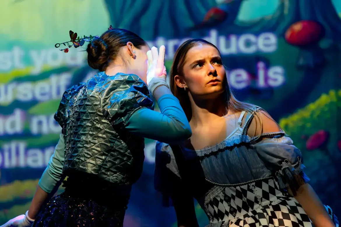 Two young women are on stage, one whispering to the other. The woman on the right has a concerned expression, wearing a costume with geometric patterns. The background shows colorful, blurred text and scenery.